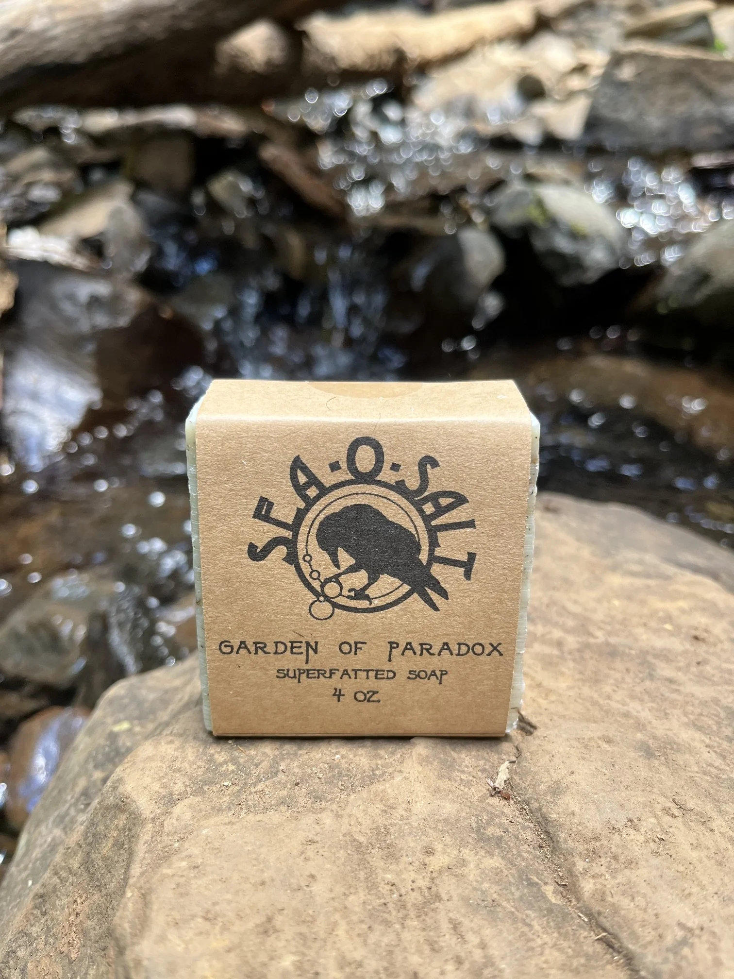 A bar of soap labeled 'Garden of Paradox Superfatted Soap 4 oz' held in front of a rocky stream with sunlight reflecting on the water.