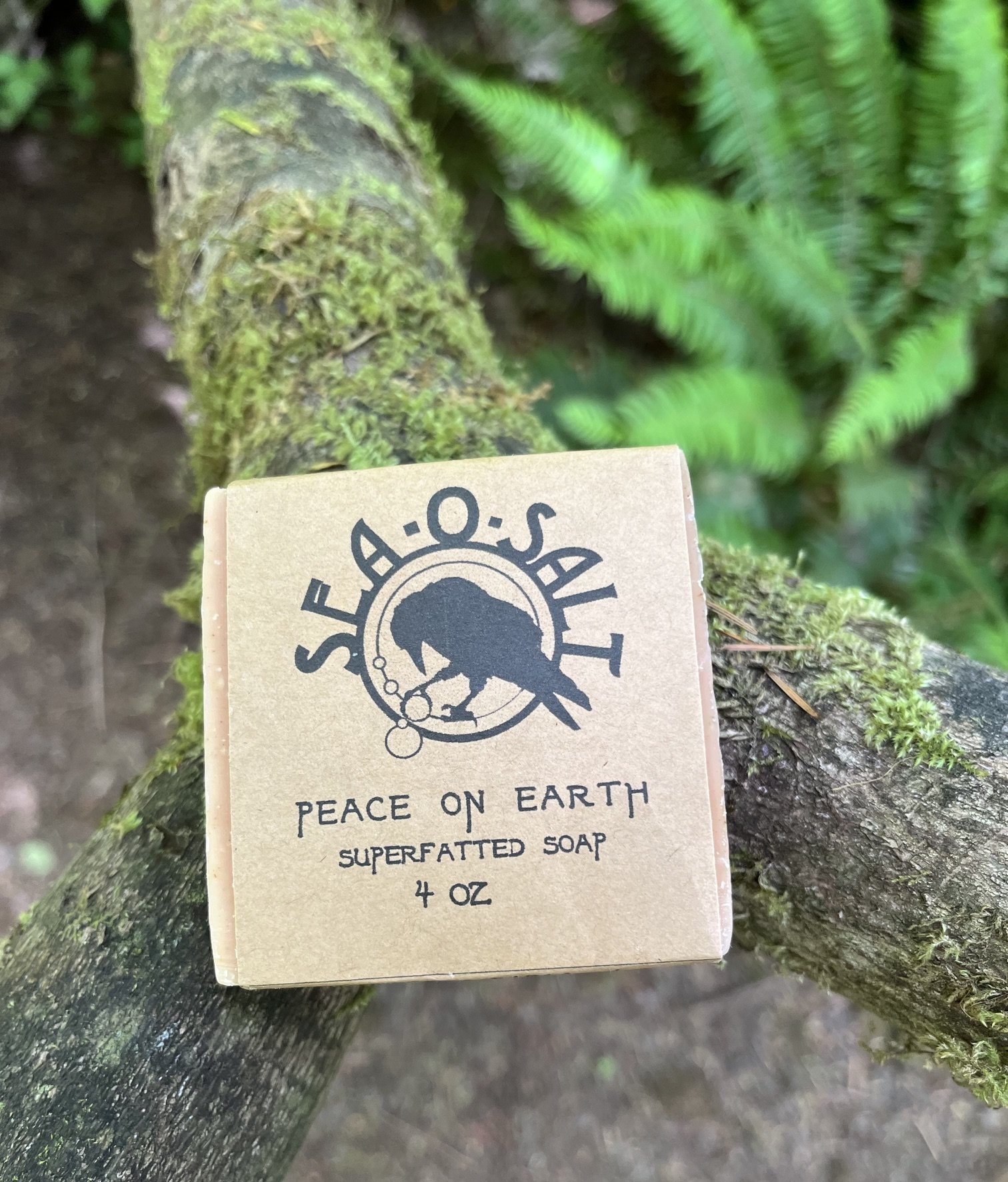 A beige label for Peace on Earth superfatting soap, placed on a moss-covered tree branch in a forest setting with ferns in the background.