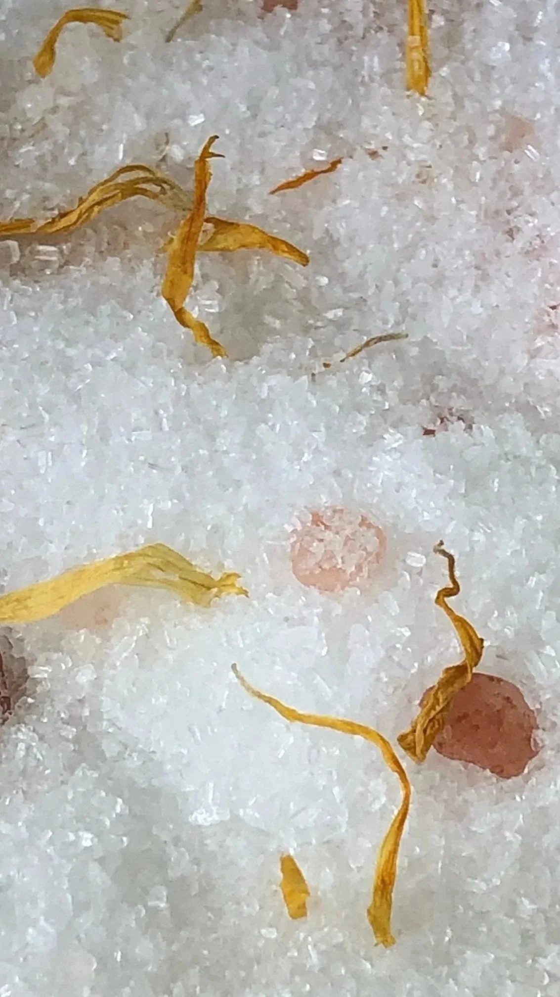 Close-up of white soaking salt with scattered yellow calendula petals and pink Himalayan salt crystals.