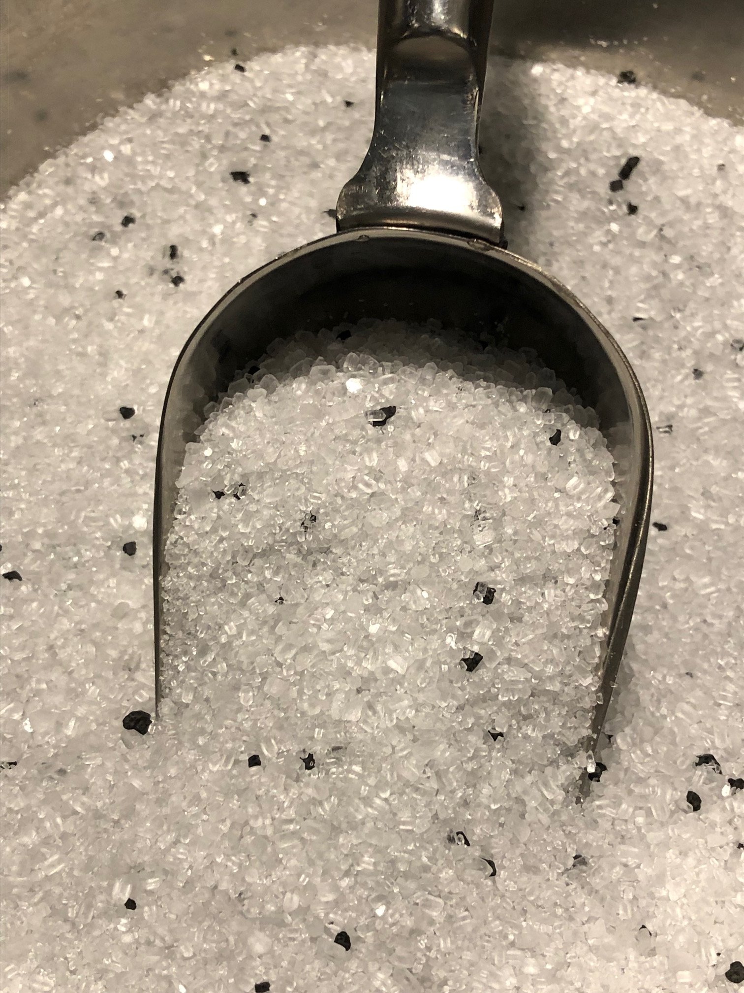 A close-up top-down view of a metal scoop digging into a bowl of soaking salt that has black Hawaiian salt crystals in it.