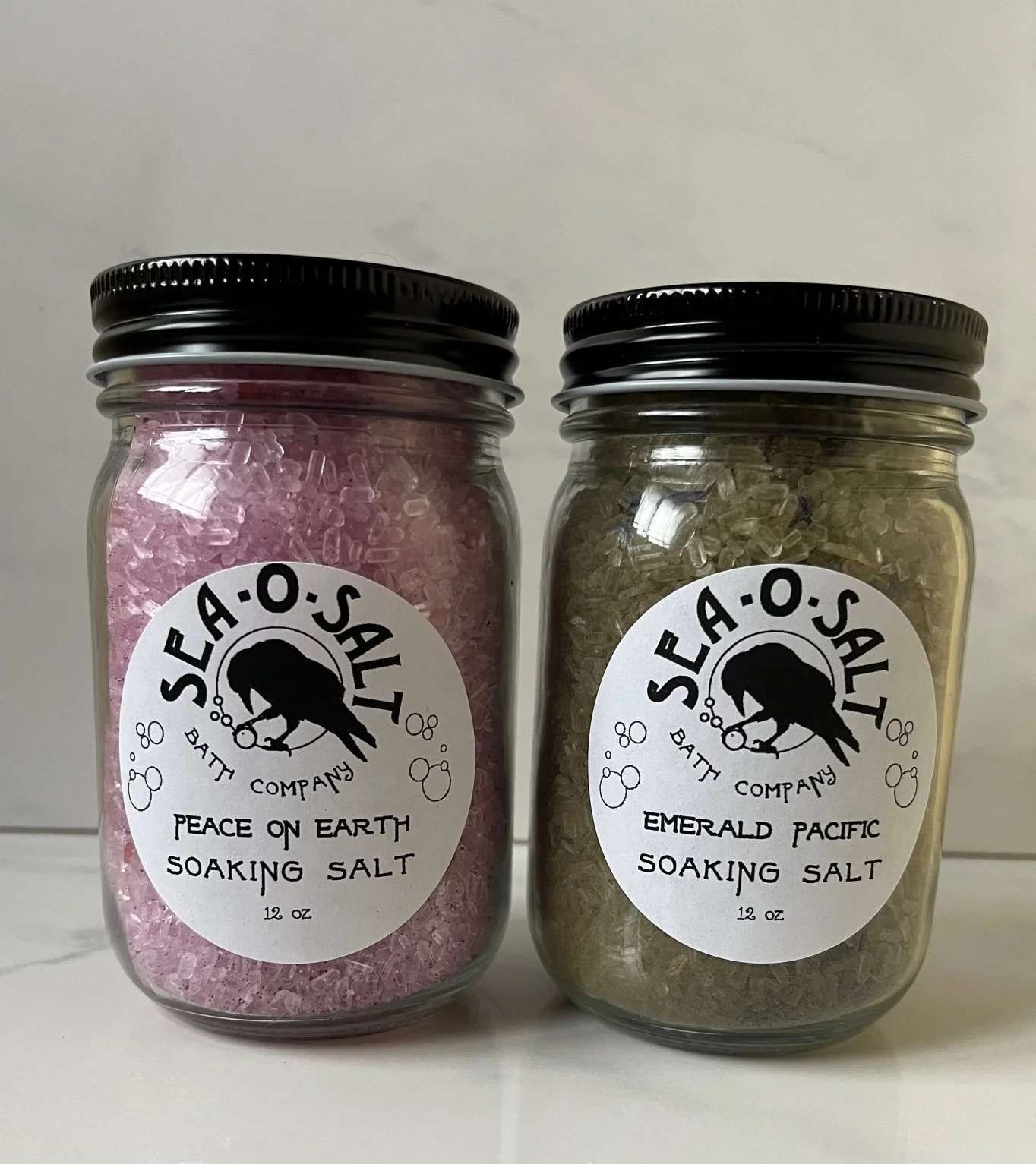 Two glass jars of soaking salts with black lids. One contains pink salt labeled 'Peace on Earth,' and the second contains green salt labeled 'Emerald Pacific.' Both jars have white labels with the Sea-O-Salt crow and bubbles logo.