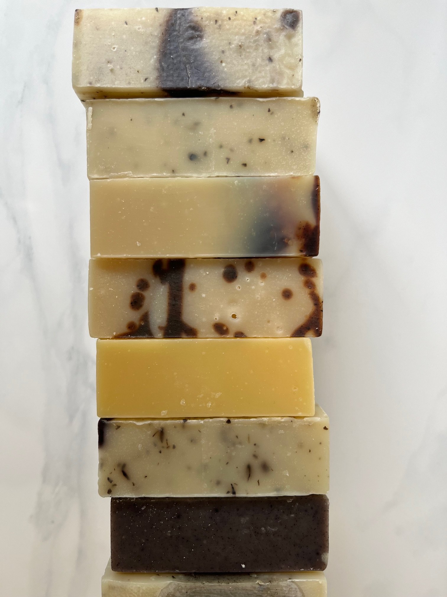 A vertical stack of ten handmade soap bars in various shades, ranging from light cream to dark brown, with some containing visible herbs and botanicals.