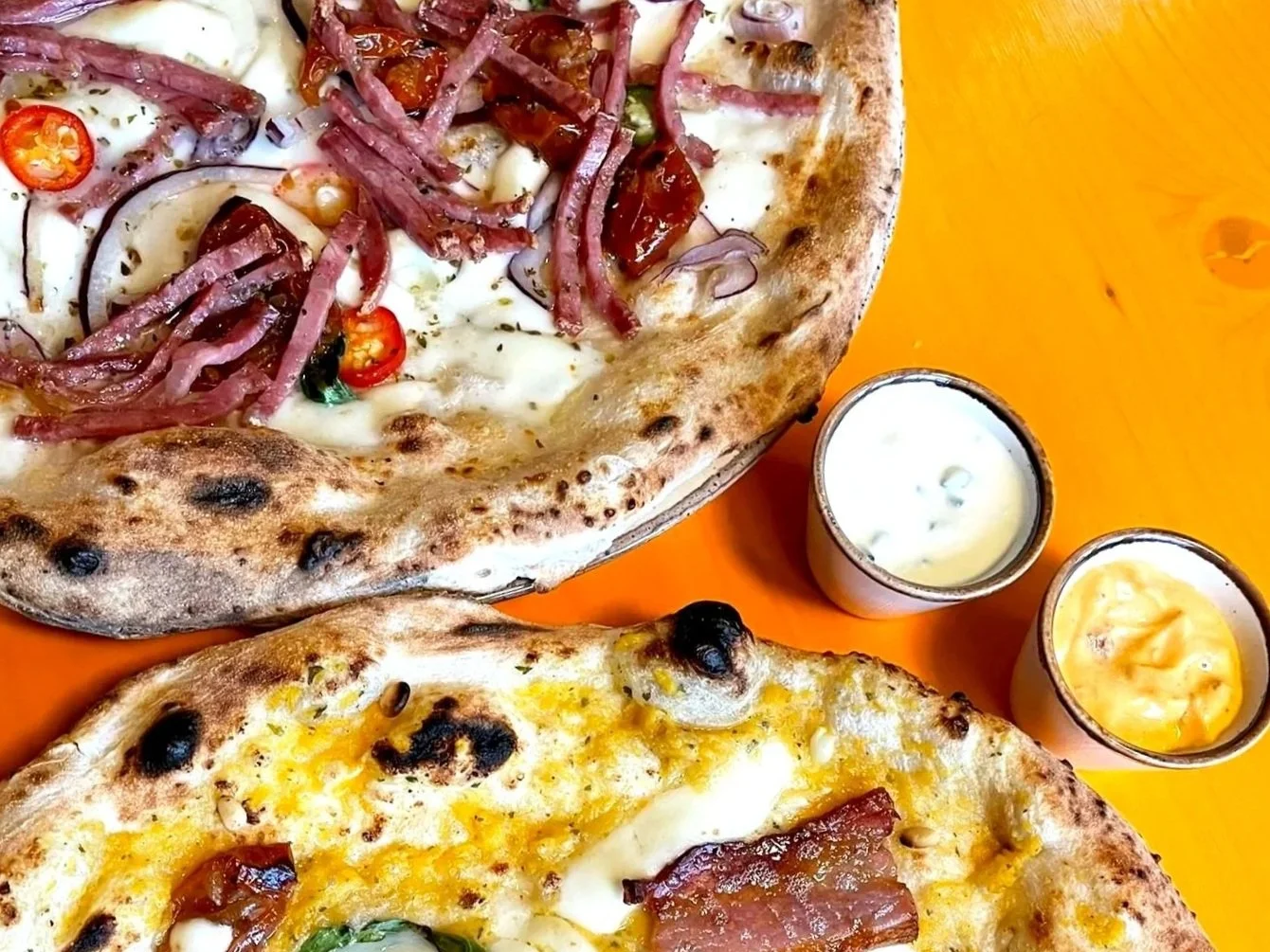 Two pizzas with various toppings, one with ham, onions, and chili peppers, and the other with cheese, ham, and green peppers. Two small cups of dipping sauces on an orange table.