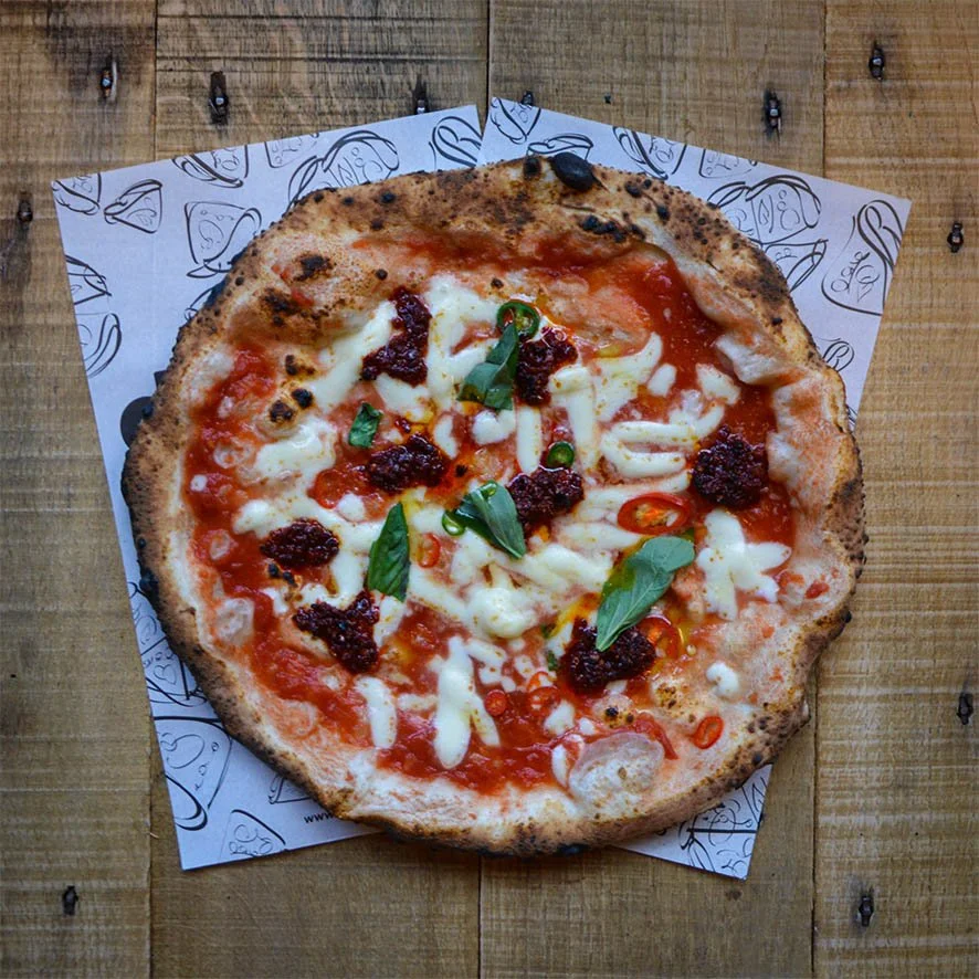 Close-up of a freshly baked pizza with cheese, tomato sauce, basil leaves, and chili slices on a wooden table, served on patterned paper.
