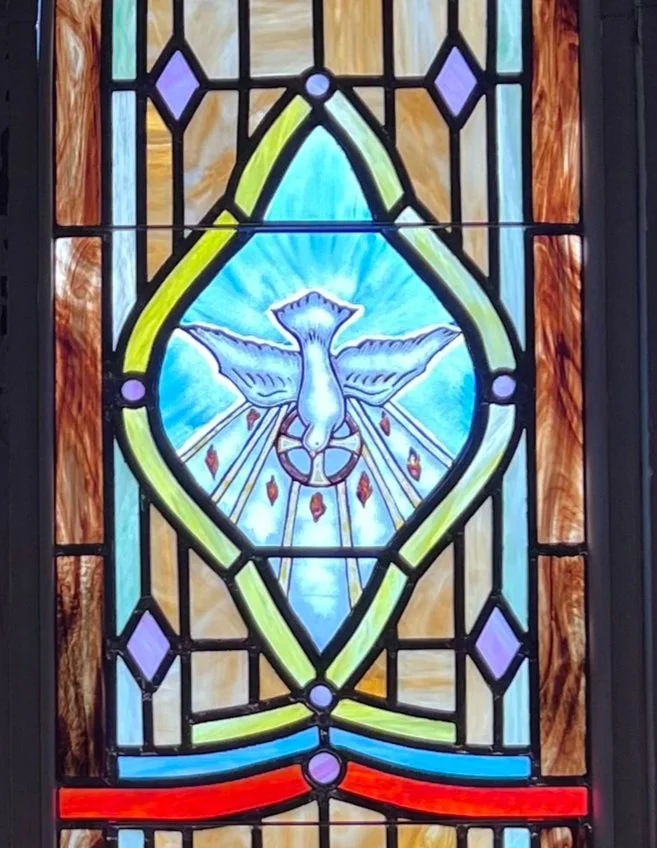 Stained glass window featuring a dove with outstretched wings, surrounded by colorful geometric patterns.