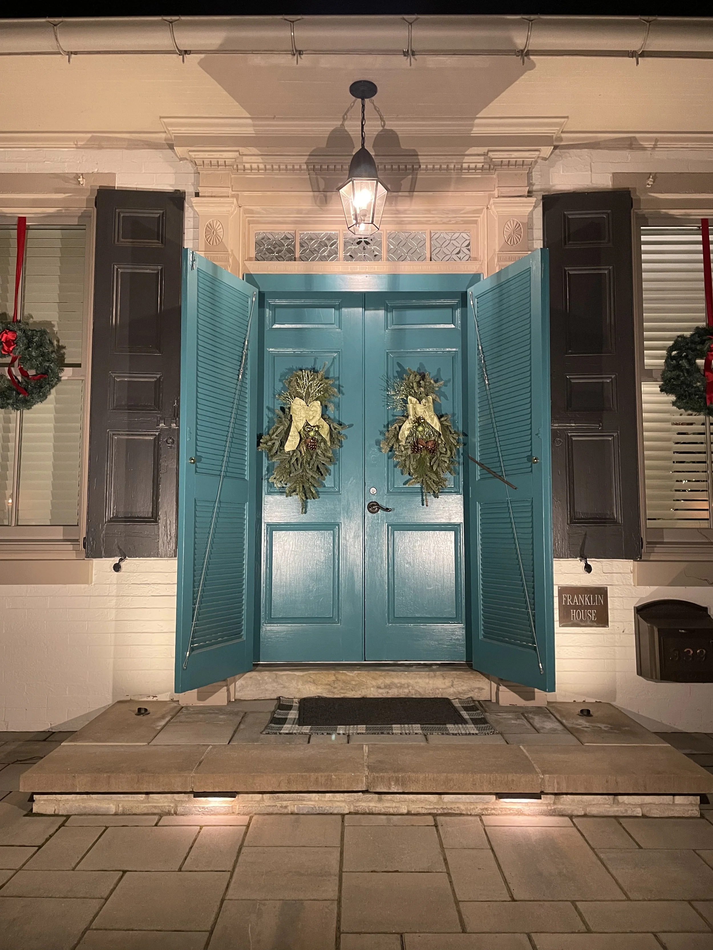 The front door of a house painted in teal with two festive wreaths hanging on the door. Flanking the door are black shutters. A porch light hangs above the door. The porch has a stone step and a black welcome mat.