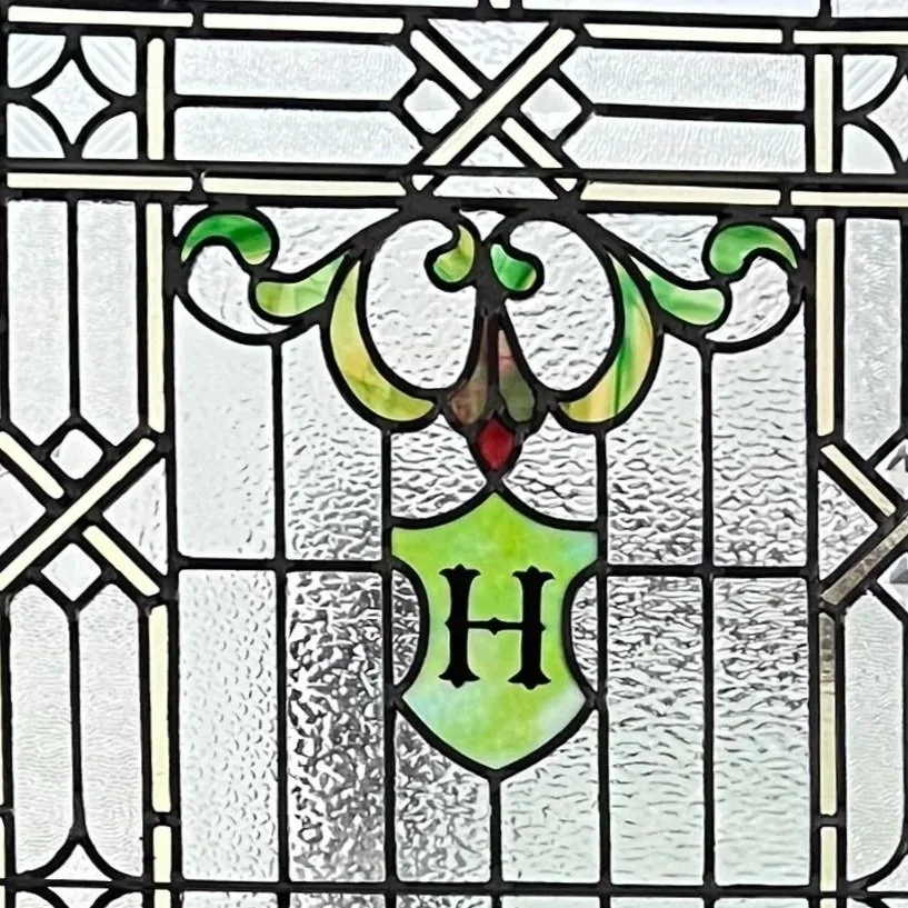Decorative stained glass window with a green shield featuring a black letter 'H' in the center, surrounded by ornate black lines and textured glass.