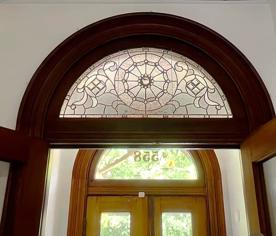 Wood-framed front door with stained glass window above, featuring intricate geometric and floral designs, with a view of greenery outside.