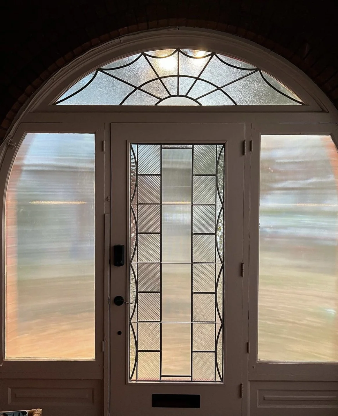 Front door with decorative glass panels and side windows, viewed from inside, with outdoor scenery blurred in the background.