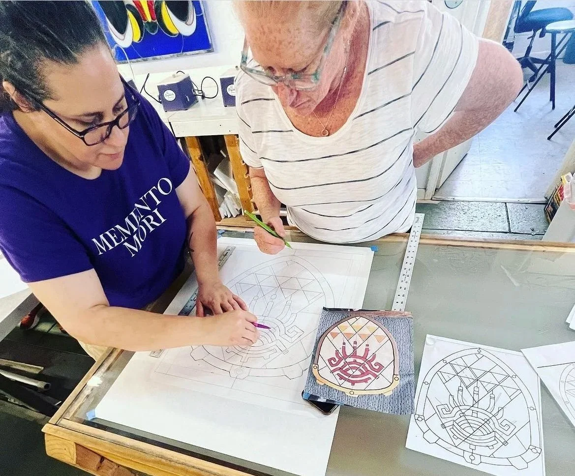 Two women are working on detailed designs of traditional textile or textile patterns at a crafting table, with sketches and a fabric piece with the pattern on it.