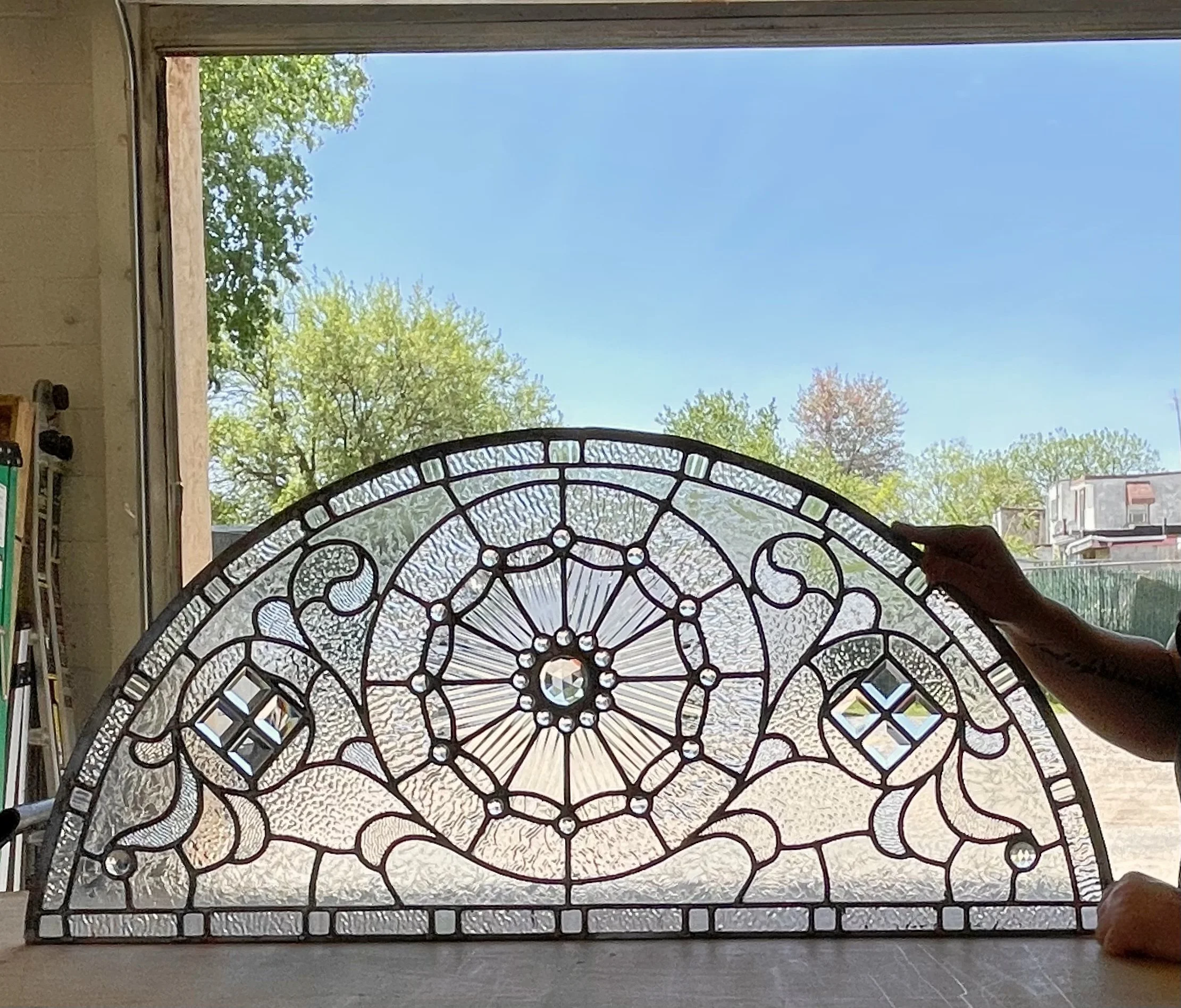 An ornate stained glass window with intricate swirling patterns, small square and circular details, and a central crystal, held by a person with an outdoor scene of trees and houses in the background visible through an open window.