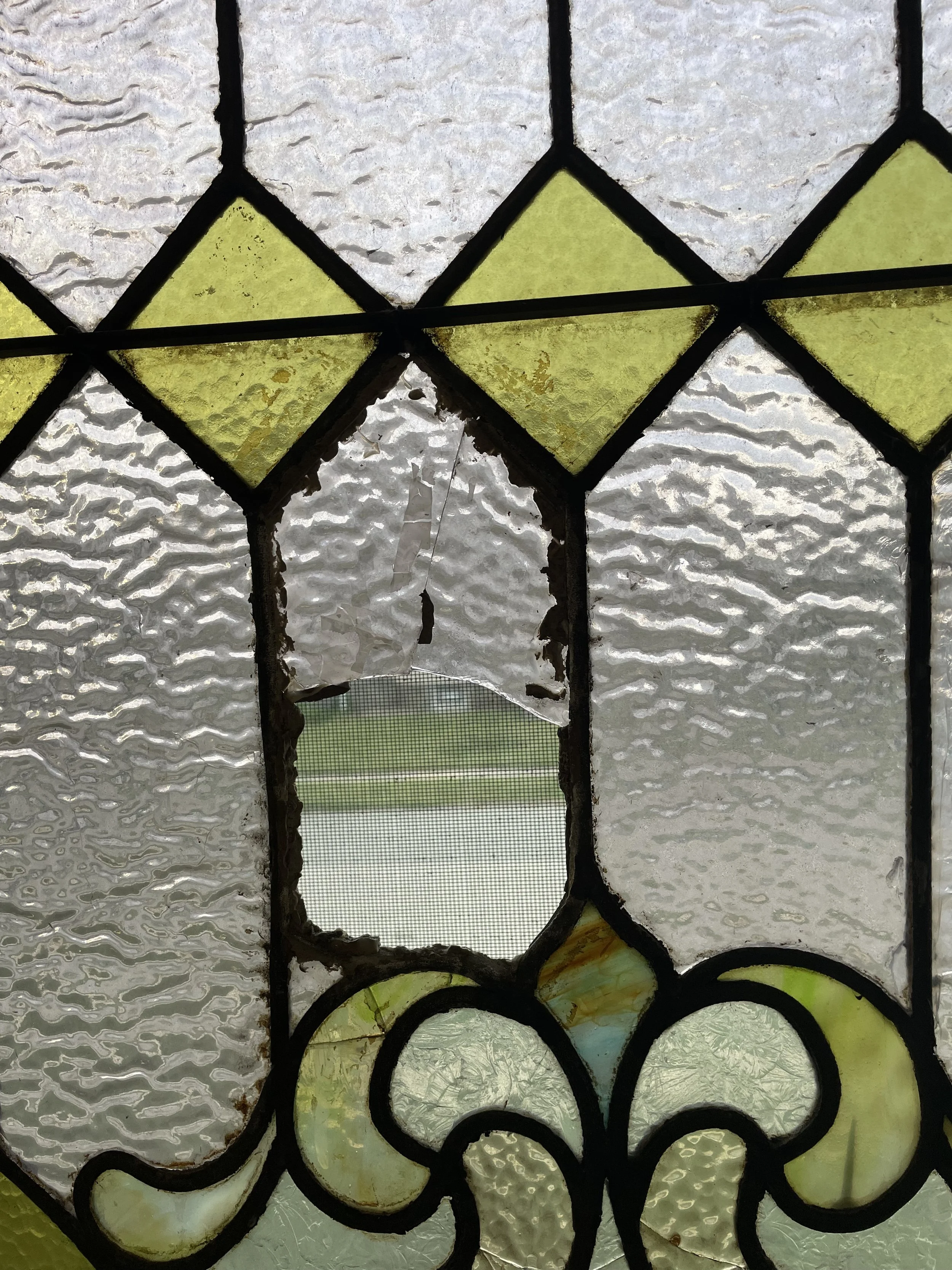 Close-up of a stained glass window with textured and colored glass pieces, some cracked and peeling, framing a view of a grassy outdoor field.