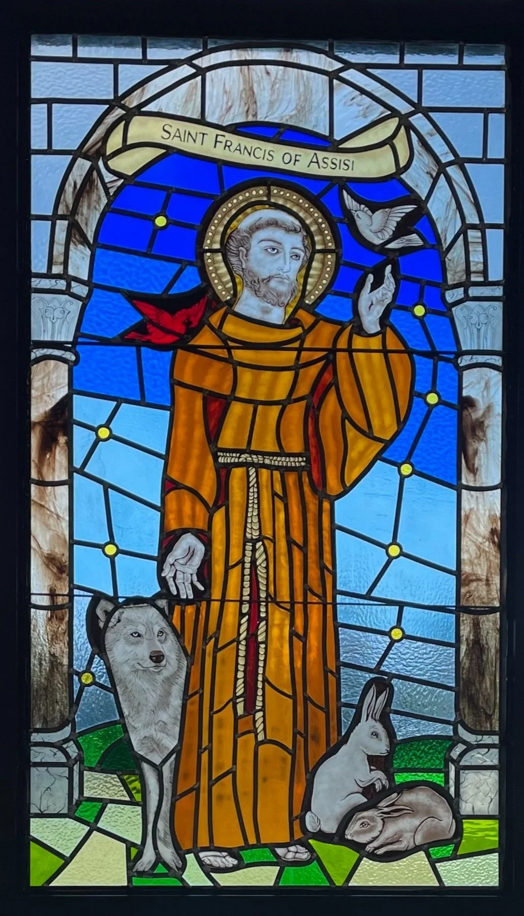 Stained glass window depicting Saint Francis of Assisi standing outdoors with animals, including a dog, a rabbit, and a bird, under a blue sky. A banner above him reads "Saint Francis of Assisi."