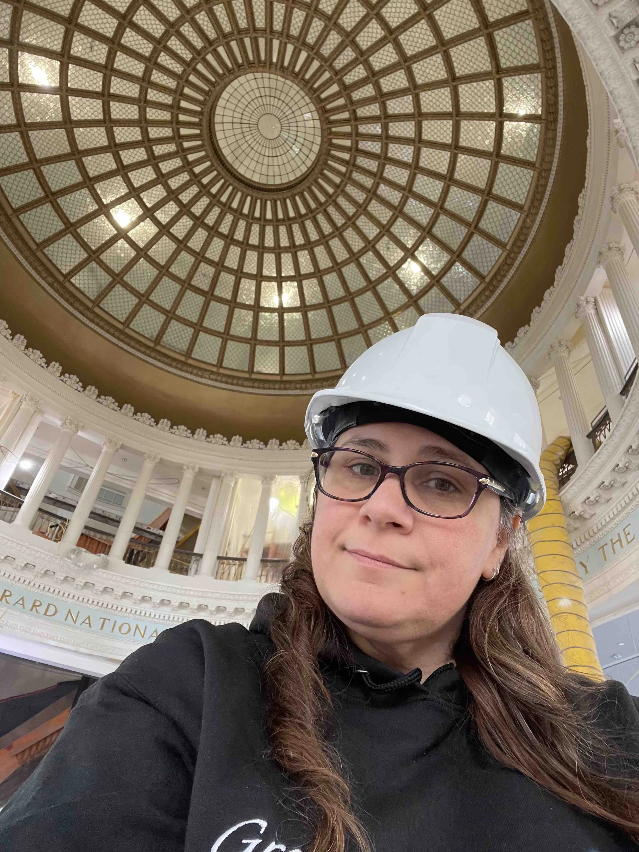 Self-portrait of a woman wearing safety glasses and a hard hat inside a grand building with a large ornate glass dome ceiling and classical columns.
