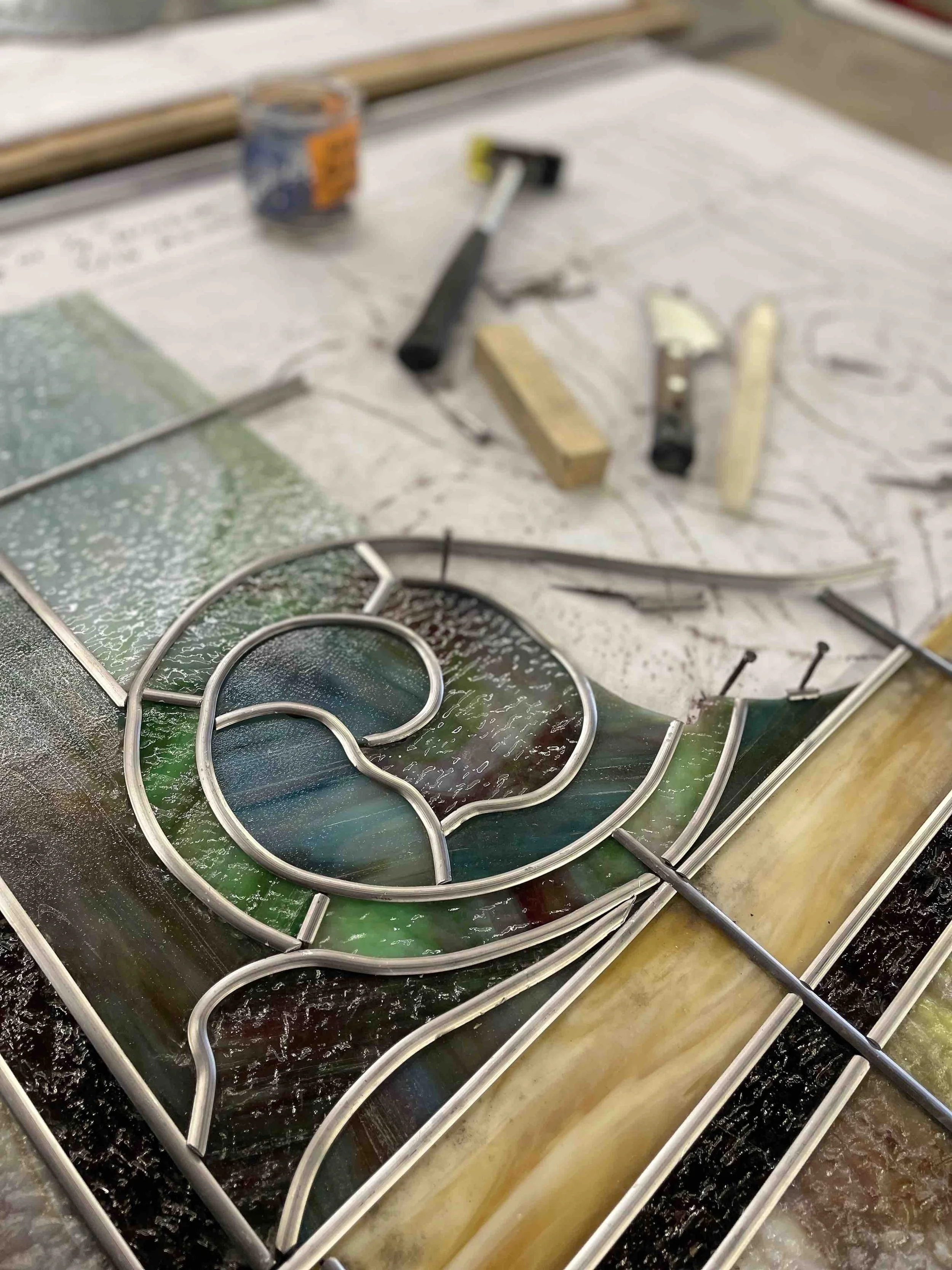Close-up of a stained glass art piece in progress, with colorful glass panels and metal framework, surrounded by tools and supplies