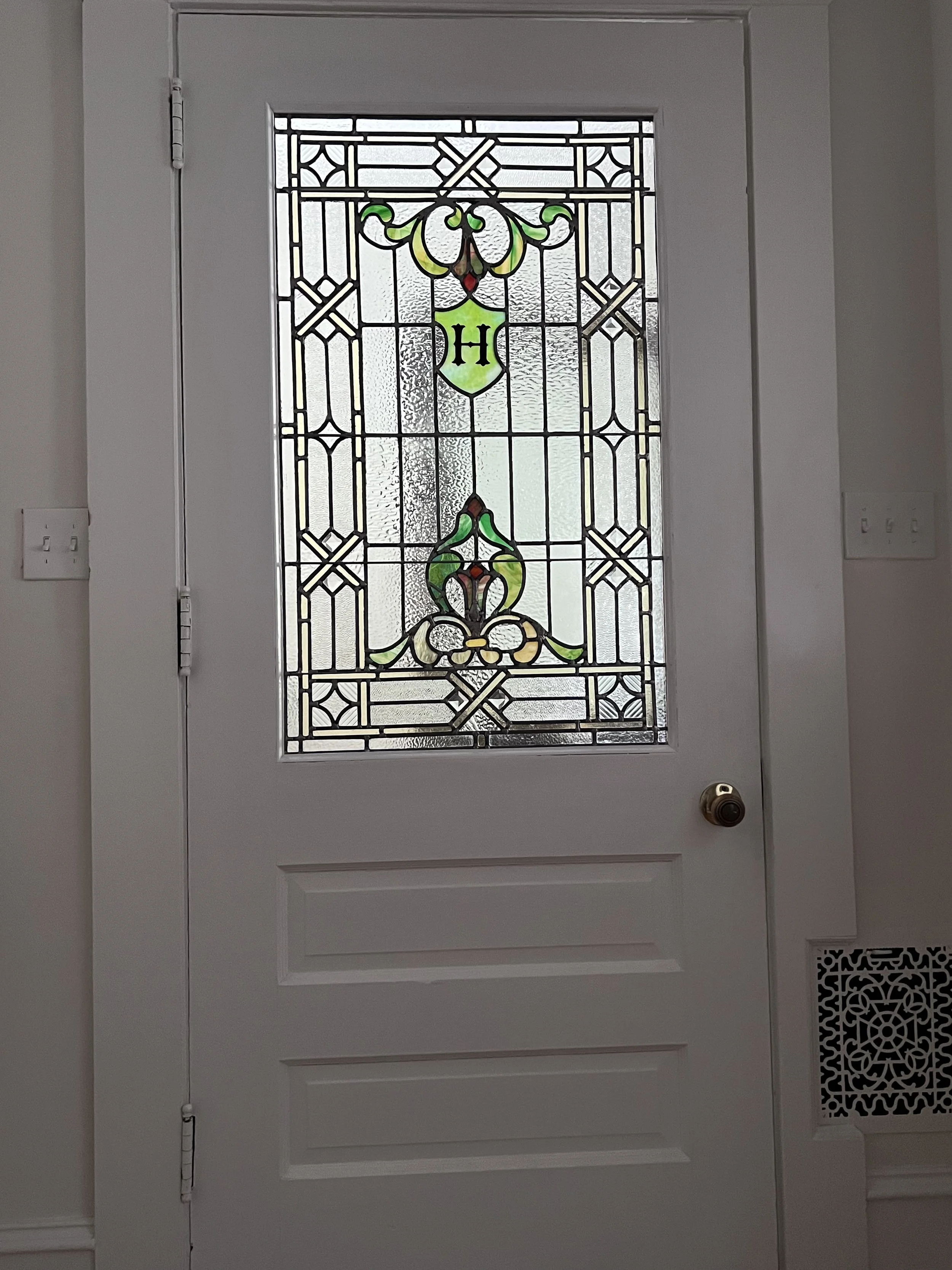 A white door with a stained glass window featuring decorative green, yellow, red, and clear glass designs, with the letter 'H' in the center.