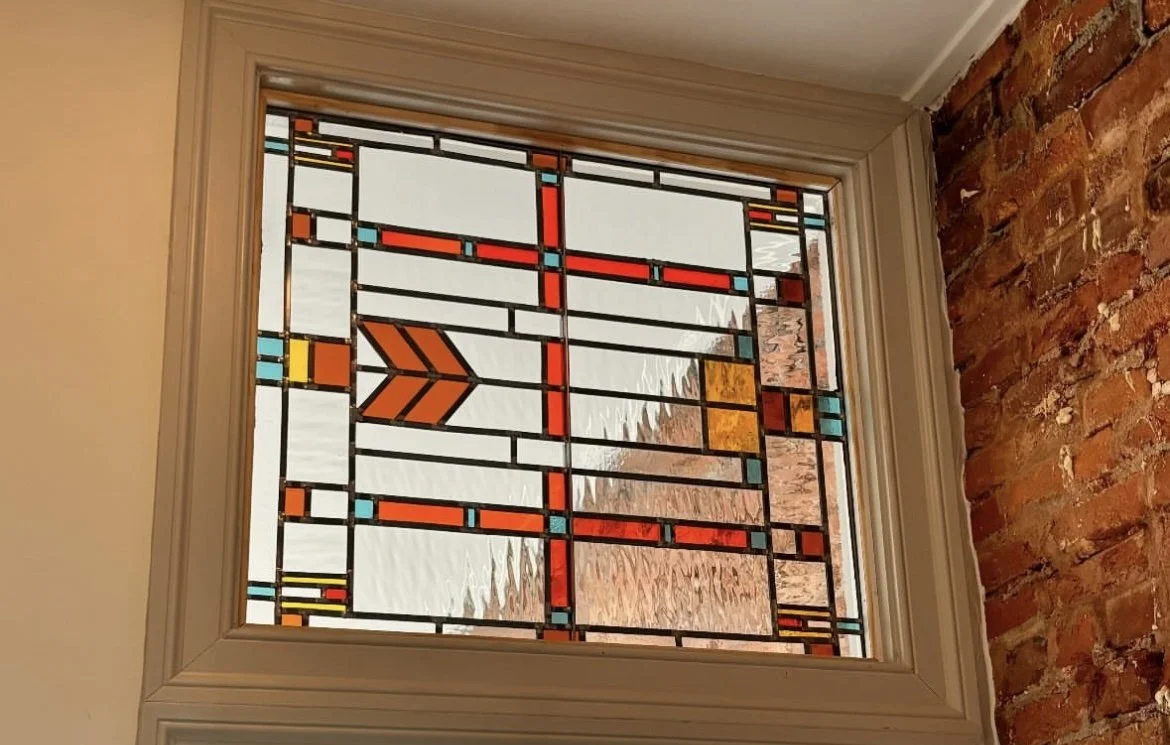 Colorful stained glass window with geometric patterns and a brick wall to the right.