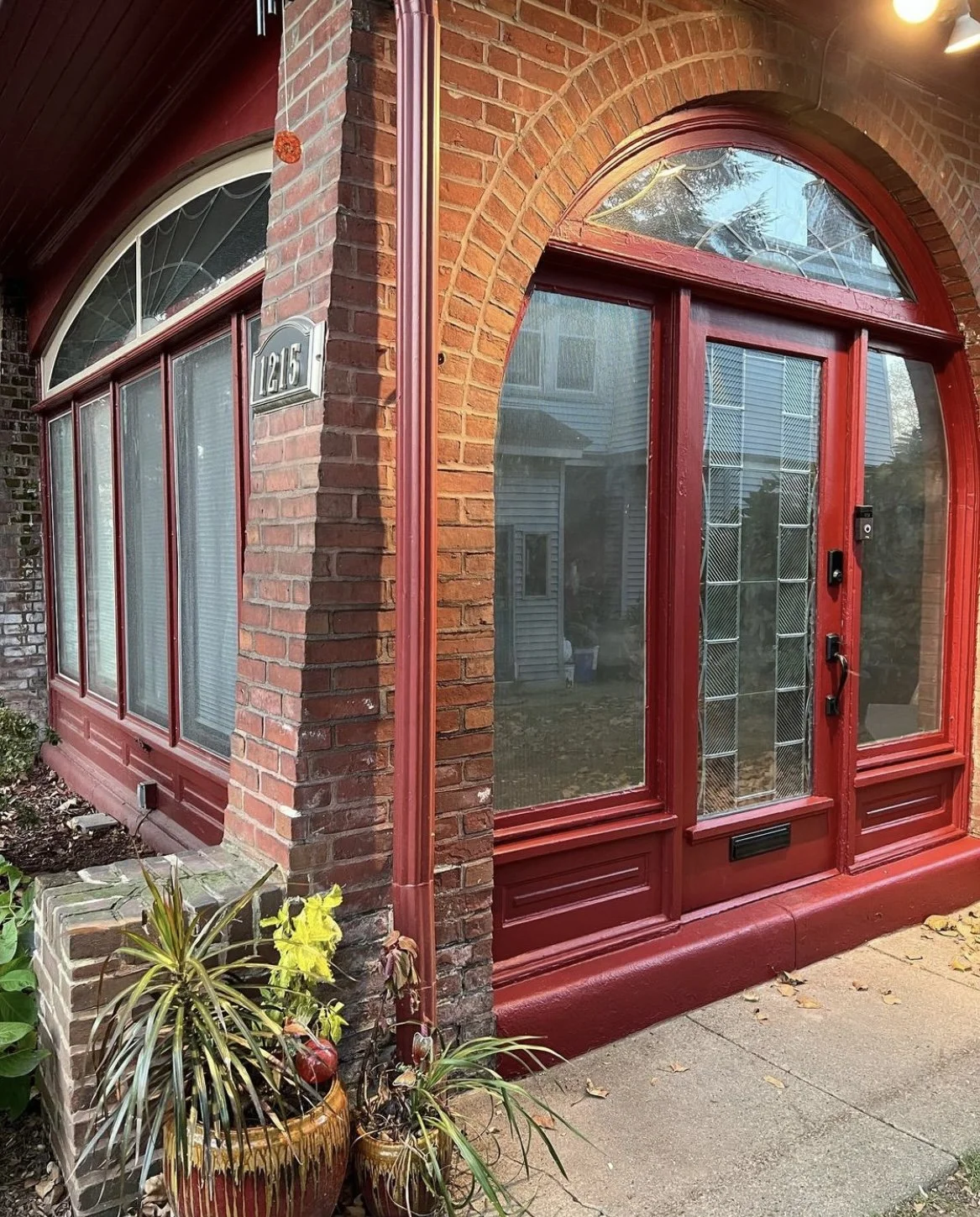 Red front door with glass panels, arched top window, brick exterior, house number 1215, and potted plants at the entrance.