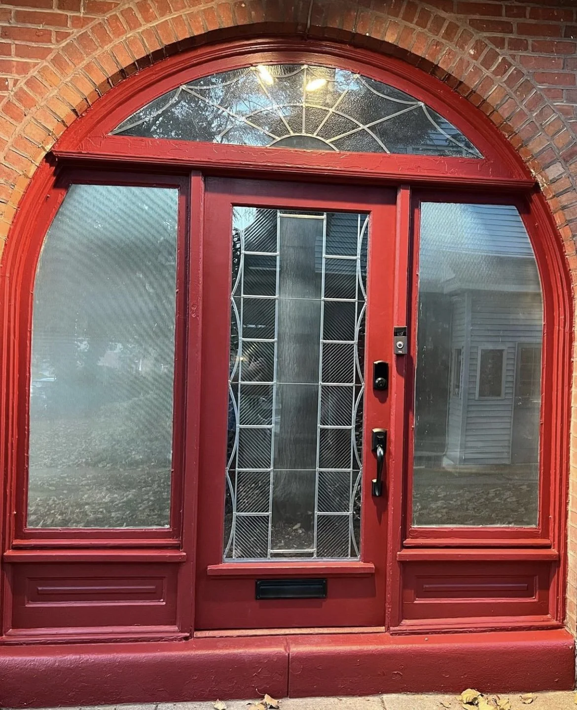Red front door with glass panels in a brick house, featuring decorative glass at the top arch and a vertical stained glass panel in the center, with a doorbell and handle on the right side.