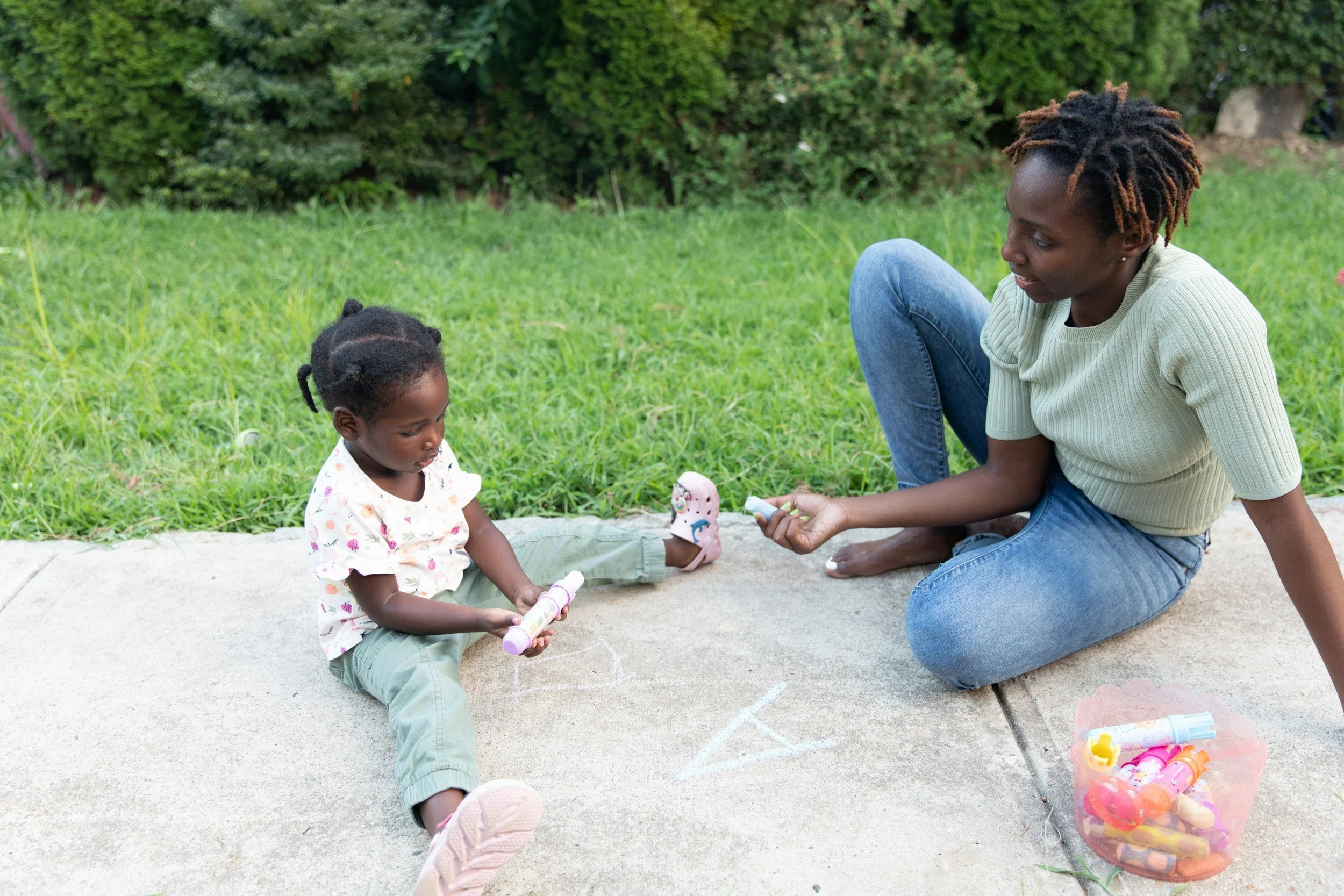 An adult woman and a young girl sitting on a sidewalk outside, playing with bubble wands. The woman is holding a bubble solution container, and the girl is holding a bubble wand. There is a plastic bag filled with colorful bubble bottles nearby. The scene is set against a grassy background and trees.