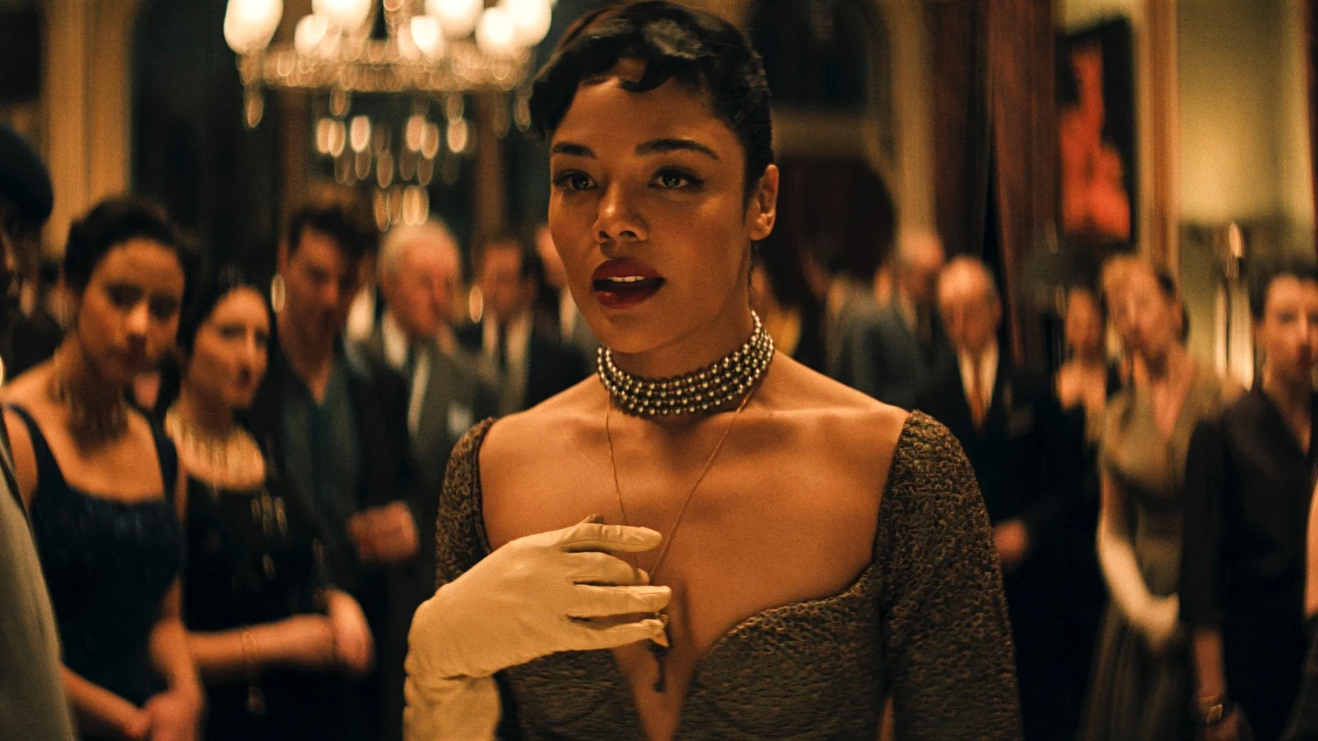 A still of Tessa Thompson in the 2025 film Hedda, standing in front of a crowd with her hand clasped to her chest.
