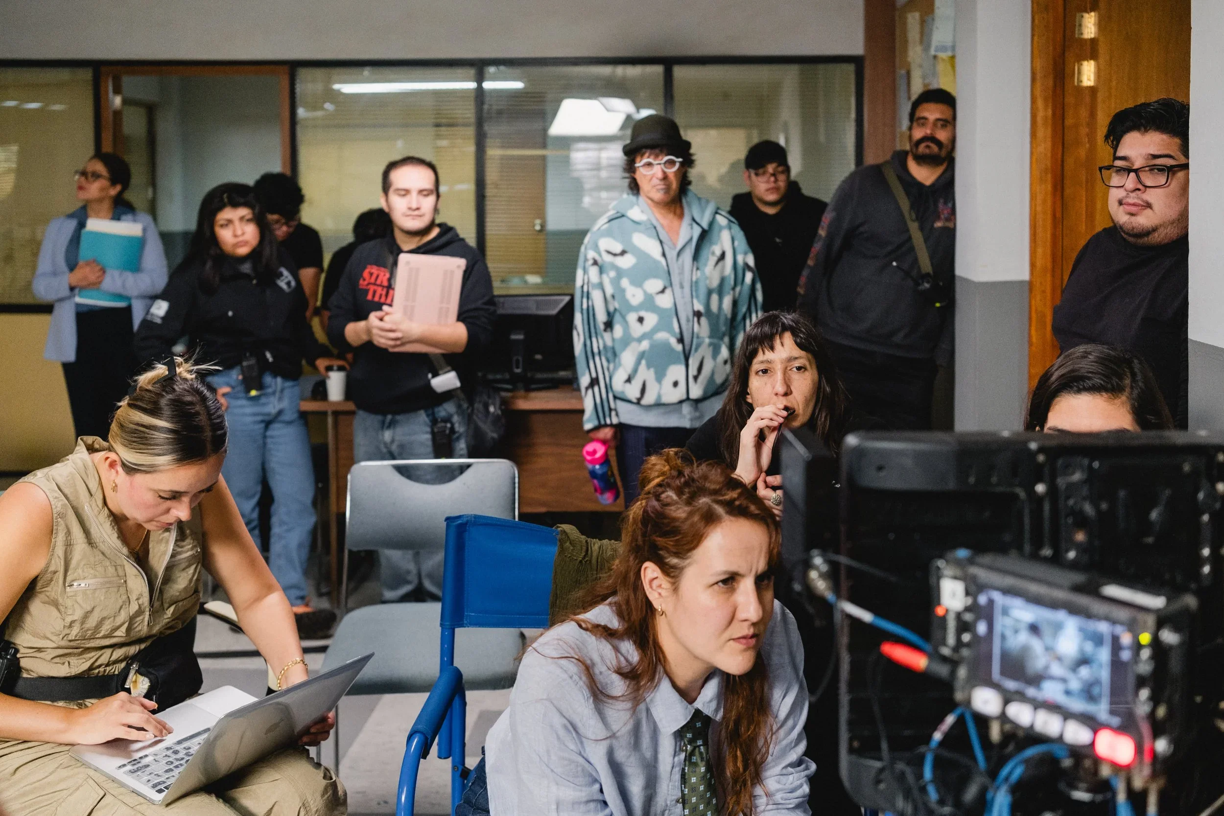 A group of people working on a film set, with some operating a camera and others in the background, in a room with a window wall.