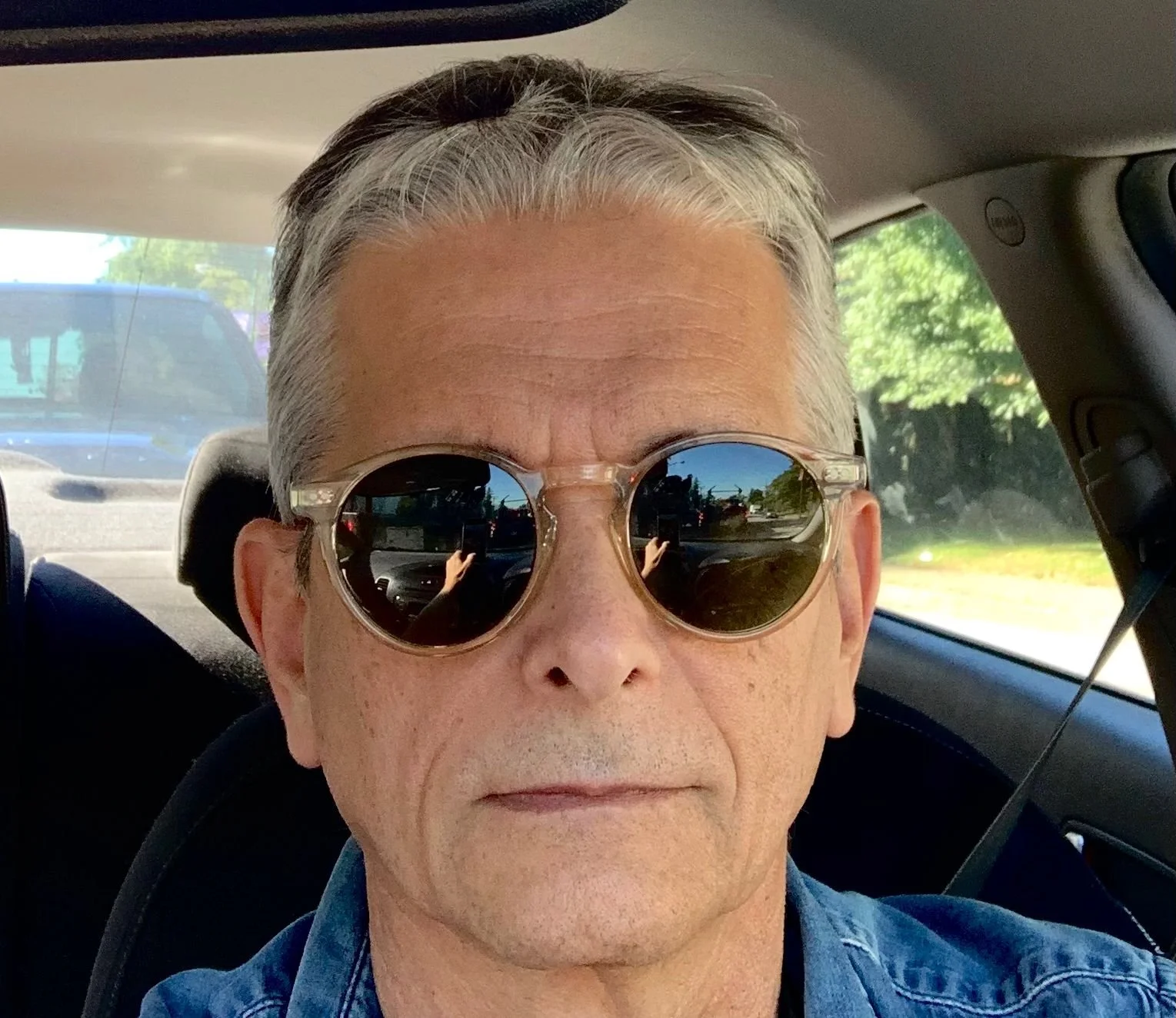 Close-up of an older man with silver hair wearing sunglasses, sitting in a car with greenery outside the window.
