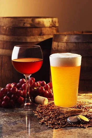 A glass of red wine, a glass of beer with foam, grapes, a cork, and a bunch of coffee beans on a wooden surface with barrels in the background. Mobile bar service.