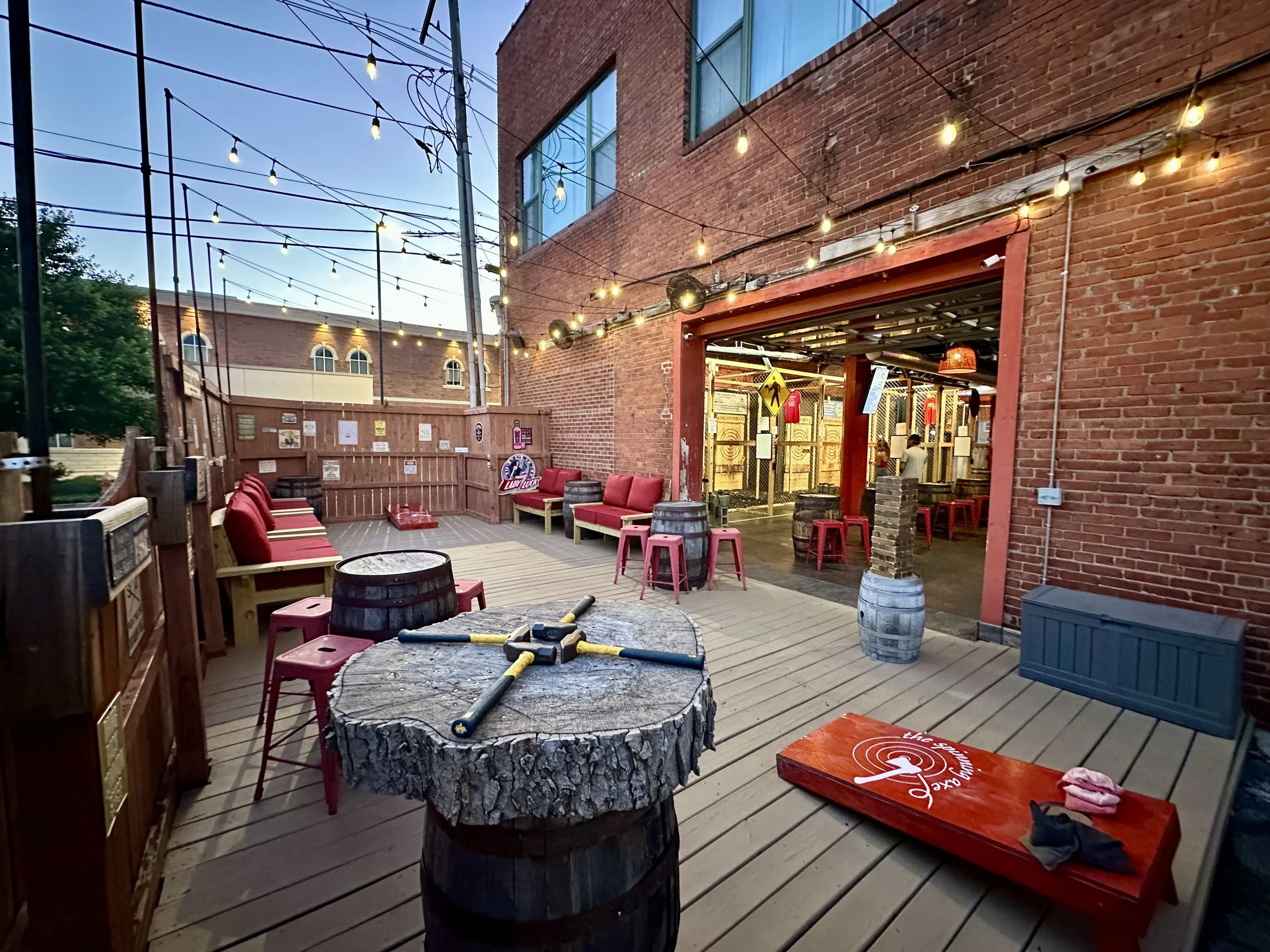 An outdoor patio area with string lights hanging above. There are rustic wooden and barrel tables, red cushioned benches, and some red stools. The scene is set against red brick walls and a building with large windows. Various signs and decorations are visible, creating a cozy, casual atmosphere.