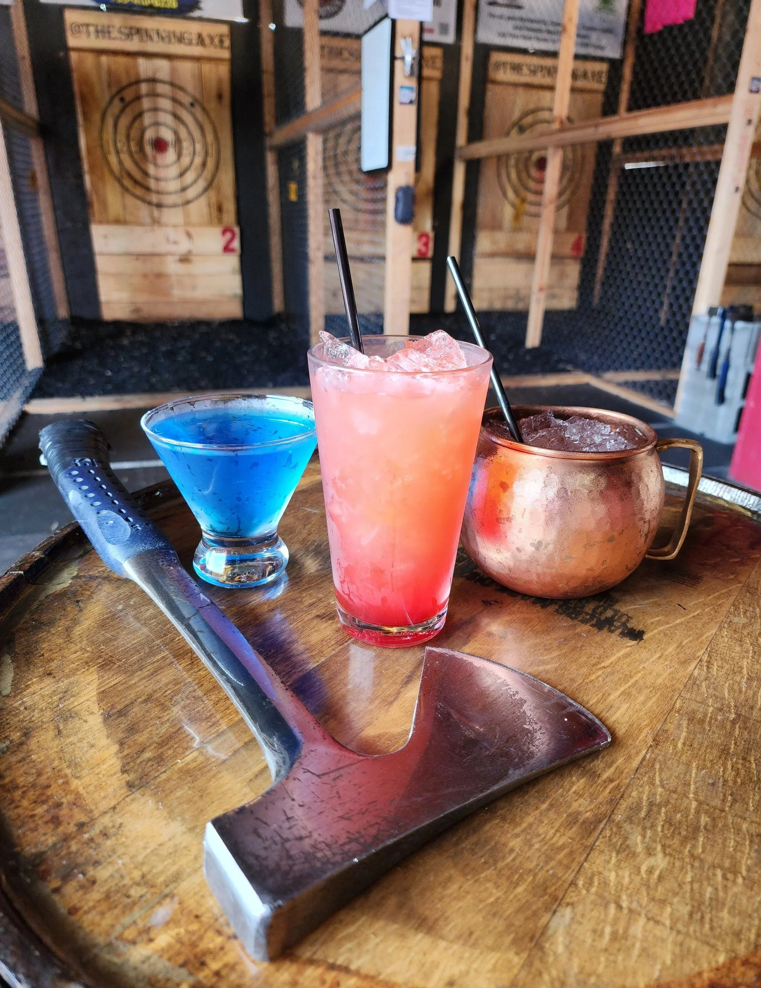 A wooden table with colorful cocktails and an axe, with targets on the wall in the background.