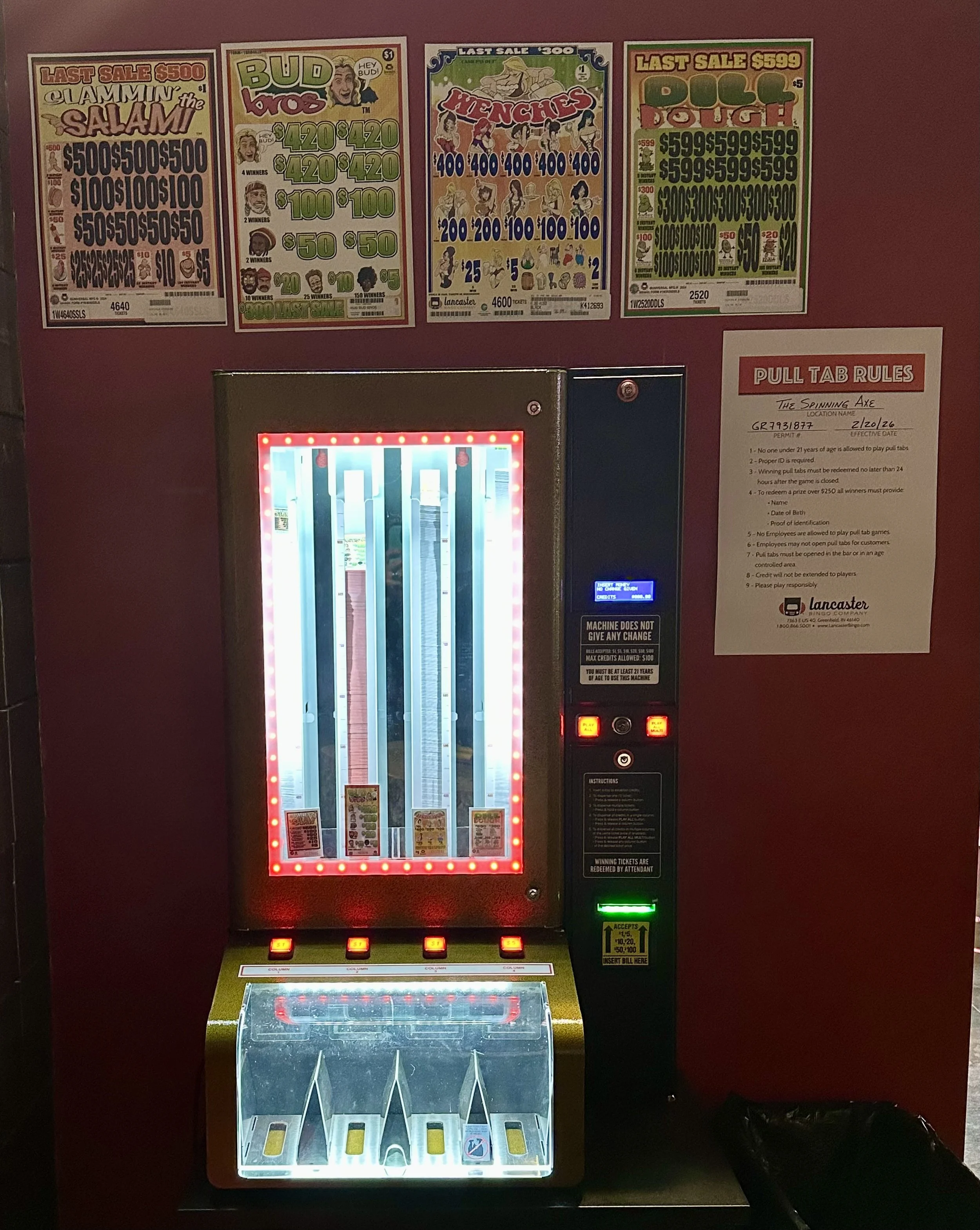 A pull tab vending machine with sale posters on the wall behind it, displaying various pull tab game prices and rules.