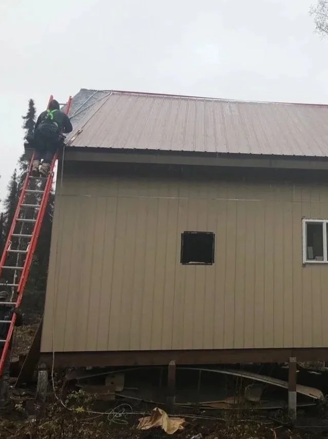 A person on a red ladder applying roofing material to the edge of a house's roof. The house has light-colored vertical siding and a small window. The scene appears to be during overcast weather.
