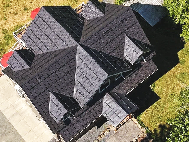 Aerial view of a house with multiple gabled roofs covered in black roofing material, surrounded by grass and trees.