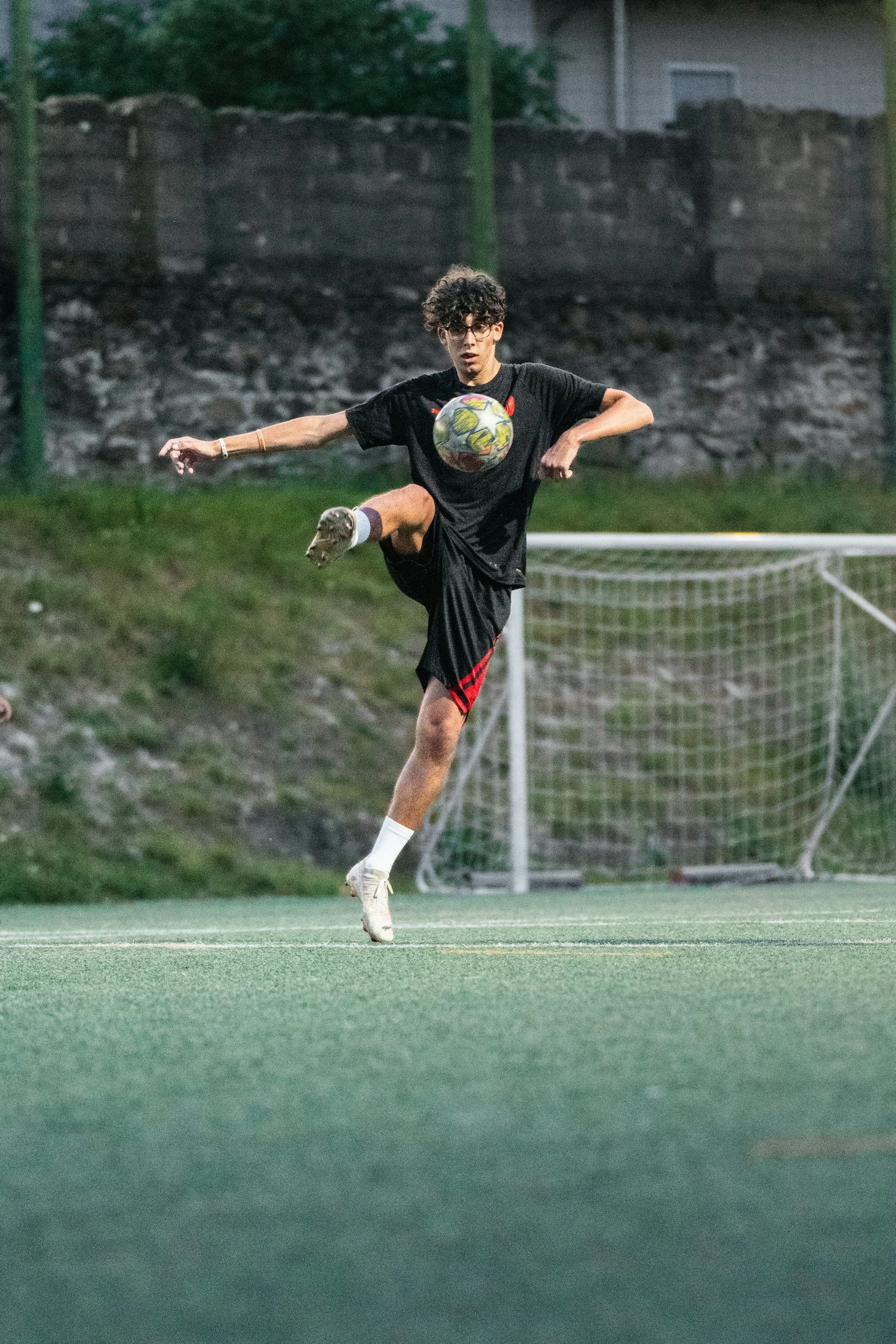 A young man playing soccer on a field, mid-action, with a soccer ball in front of him, wearing black sportswear with red stripes, white cleats, and glasses.