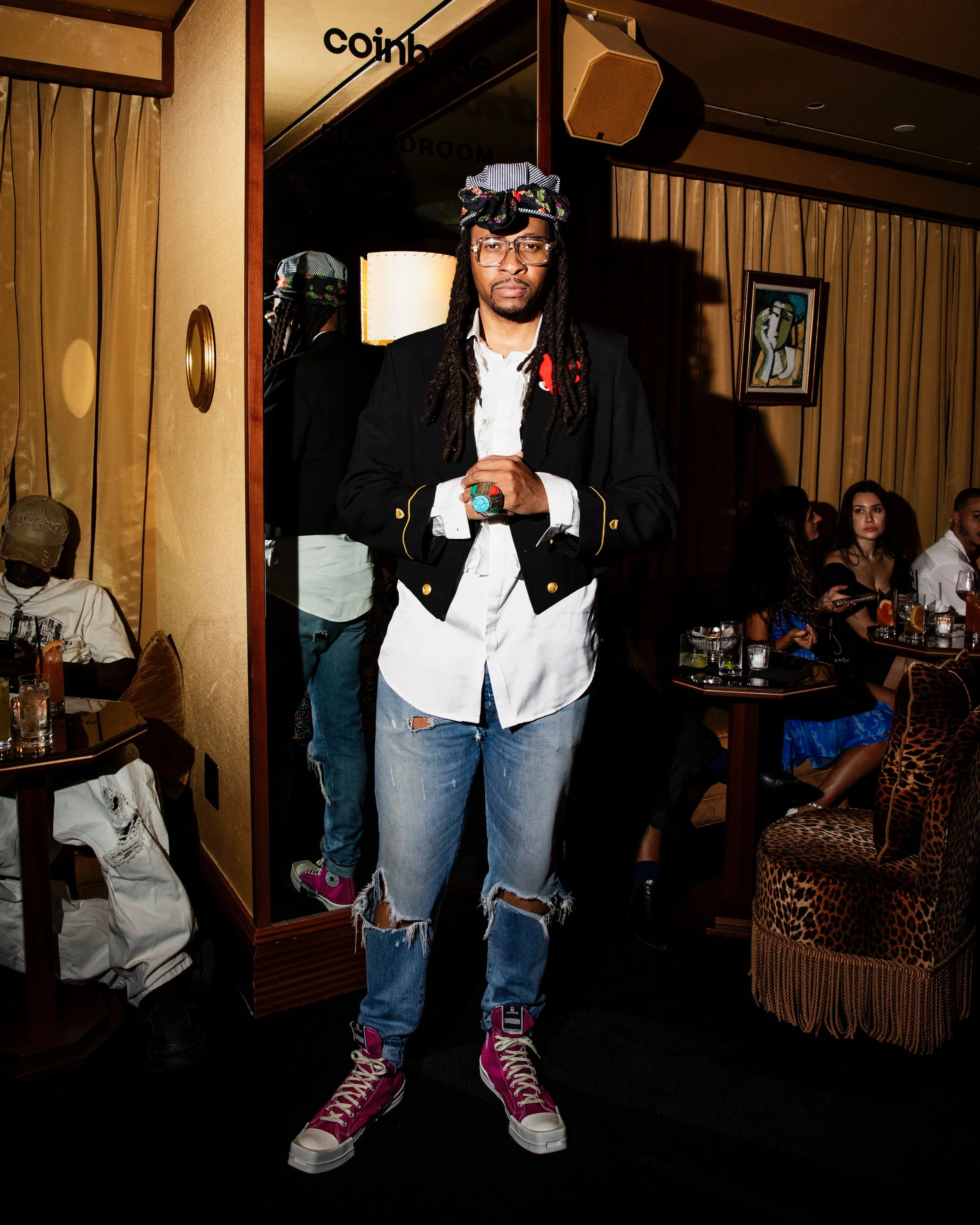 A man with glasses, dreadlocks, and a striped cap standing in a room with warm lighting. He is wearing a black blazer with gold accents over a white shirt, ripped jeans, and bright pink high-top sneakers. He has his hands clasped in front of him and 