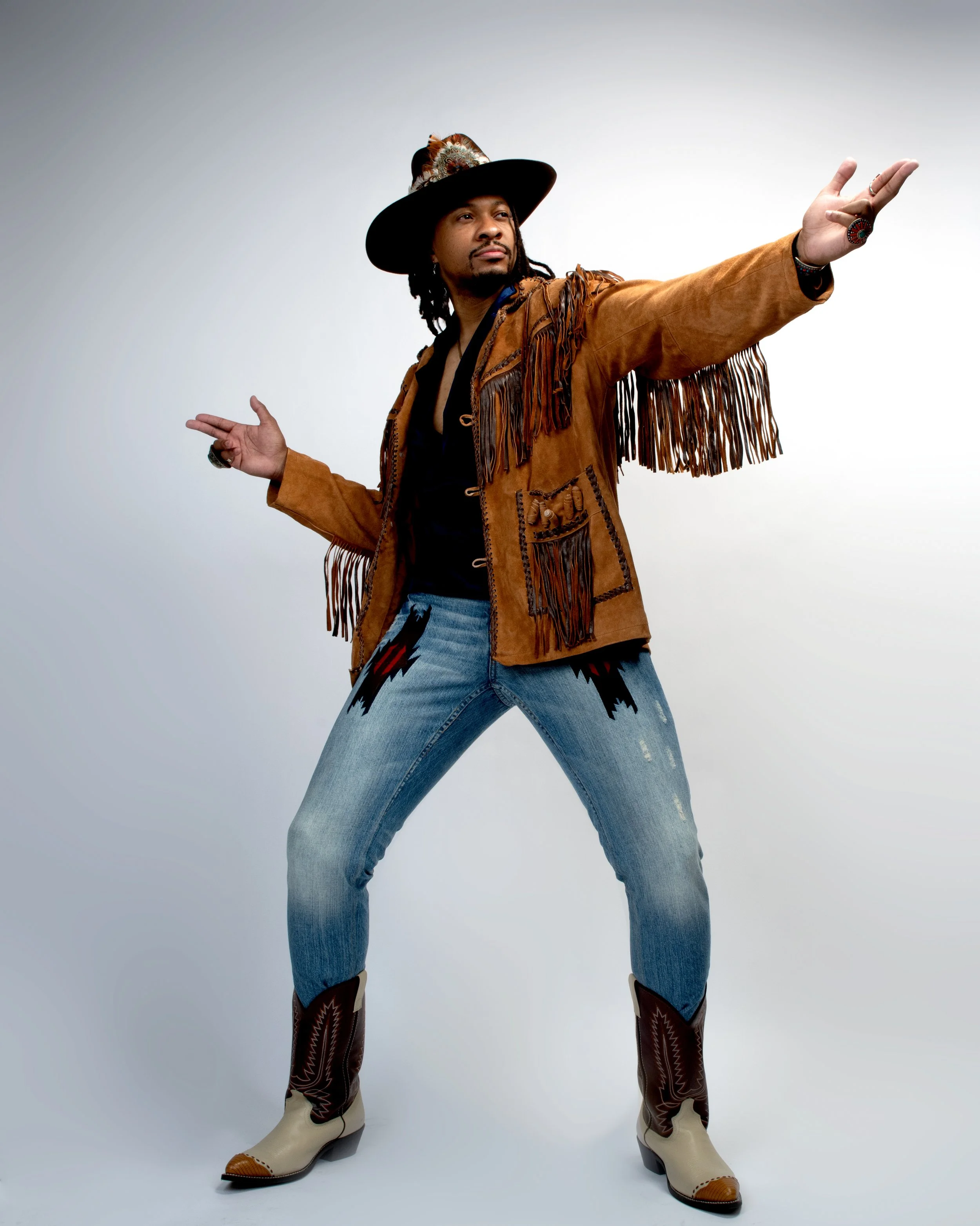 A man dressed in vintage western attire stands against a plain gray background, with one arm extended outward and the other bent, wearing a feathered hat, fringed suede jacket, patterned jeans, and cowboy boots.