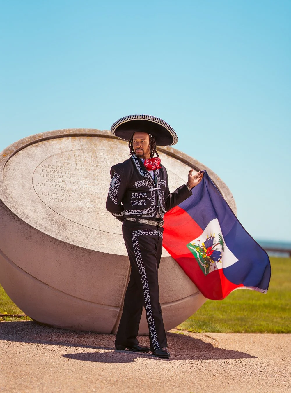 A man dressed in traditional Mexican clothing, wearing a large sombrero, holding a Haitian flag, standing in front of a stone monument with engraved text under a clear blue sky.