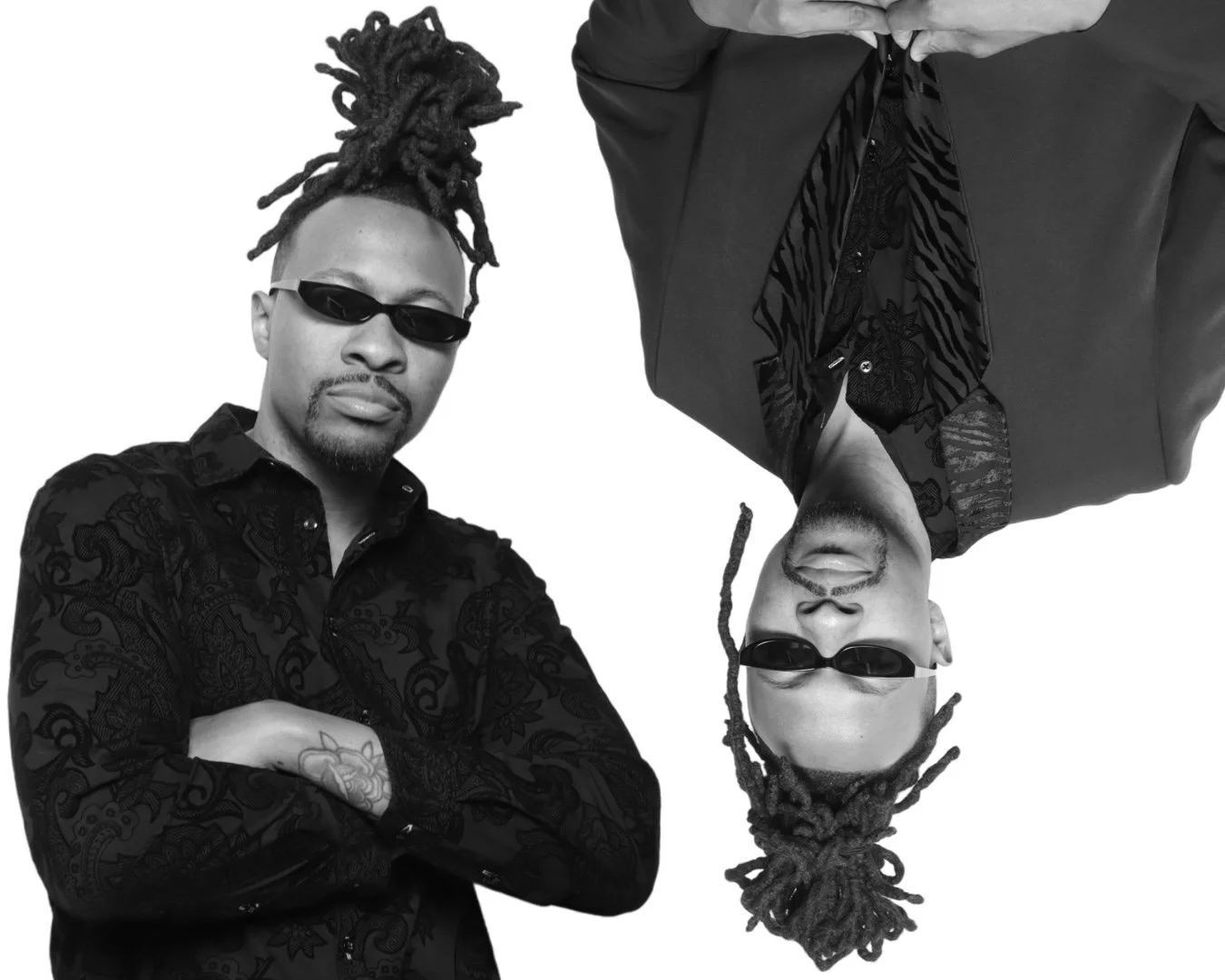 Two men wearing black clothing and sunglasses, sitting against a plain background. One has dreadlocks and the other has a shaved head. The image is in black and white.