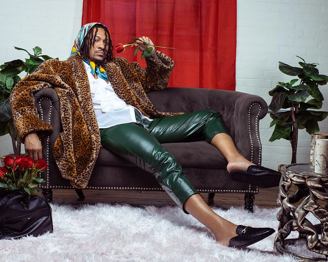 Person sitting on a sofa in a leopard print coat, green leather pants, and black loafers, holding a red rose in a room with plants and a white brick wall.