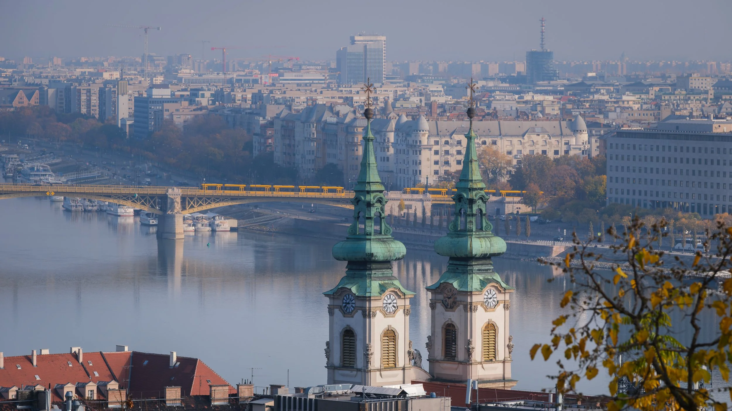 A view across the Danube through the green towers of the Church of St Anne and towards the Margaret Bridge with two yellow city trams passing by.