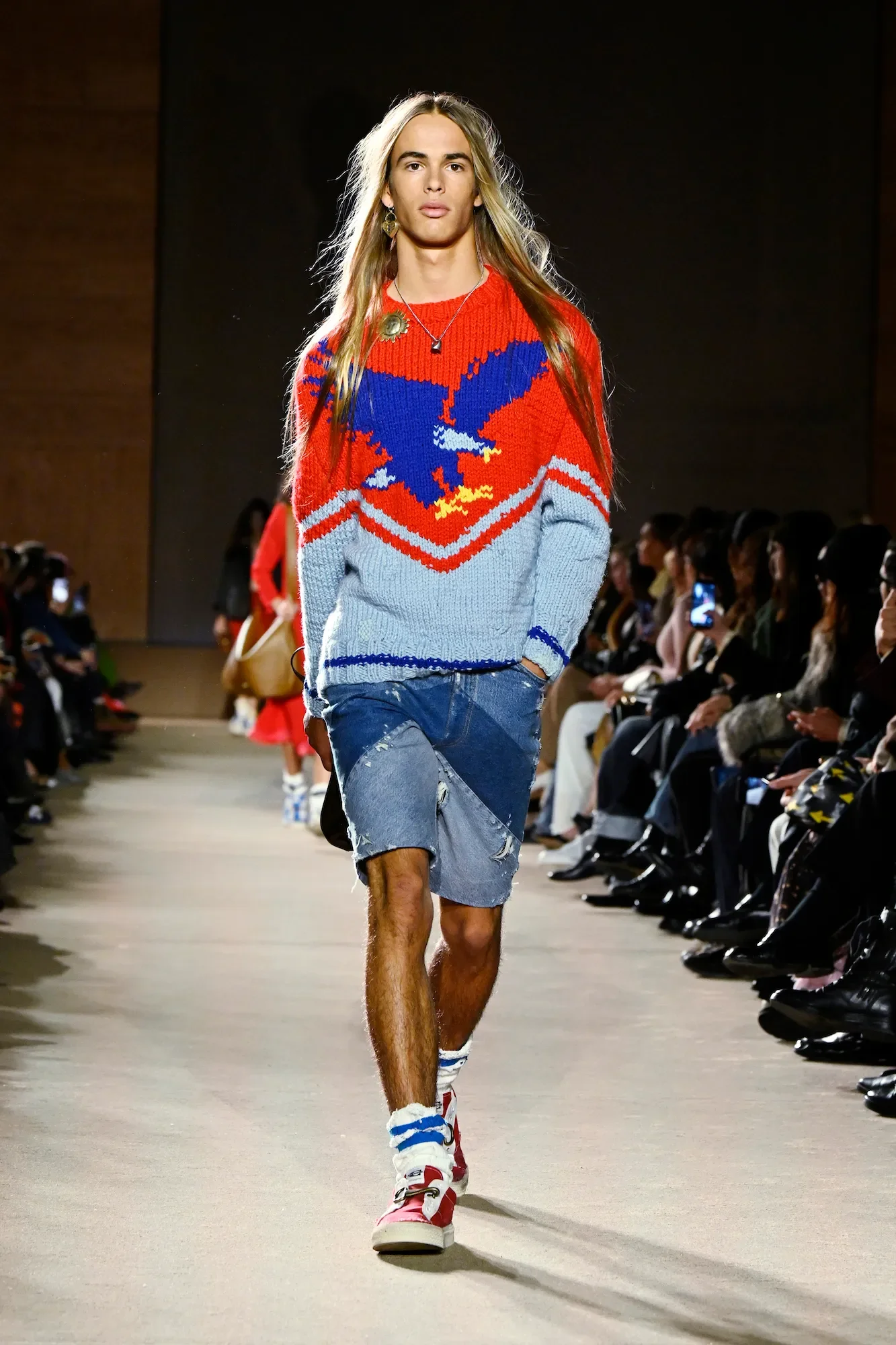 Male model walking down a runway during a fashion show, wearing a colorful sweater with an eagle design, denim shorts, and sneakers. Audience seated on both sides.