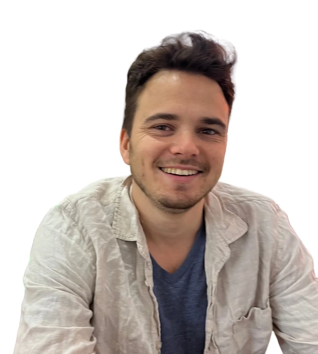 A smiling young man with dark, styled hair, wearing a beige jacket over a blue shirt, against a plain white background.