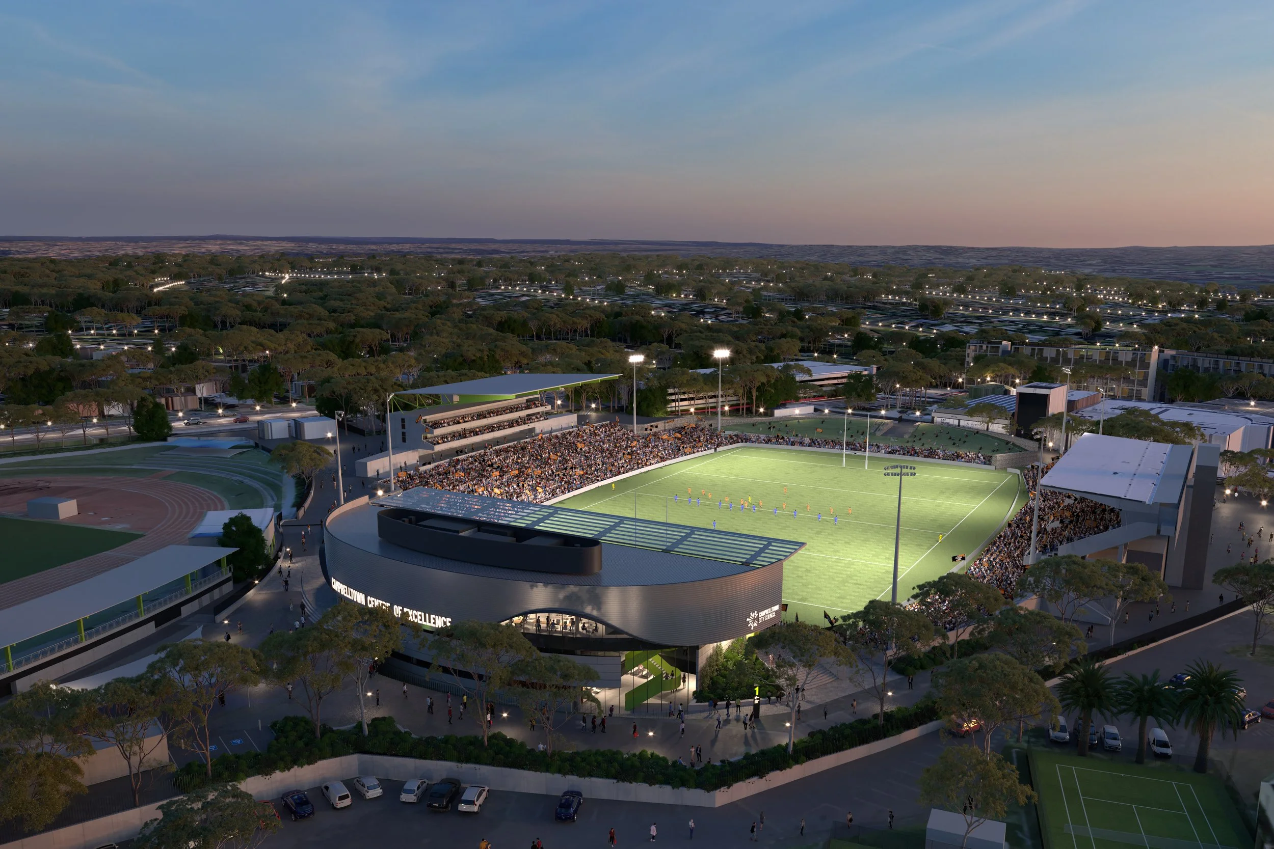 Aerial view of a sports stadium at dusk, filled with spectators, with a football game in progress on the field, surrounded by trees and a parking lot.