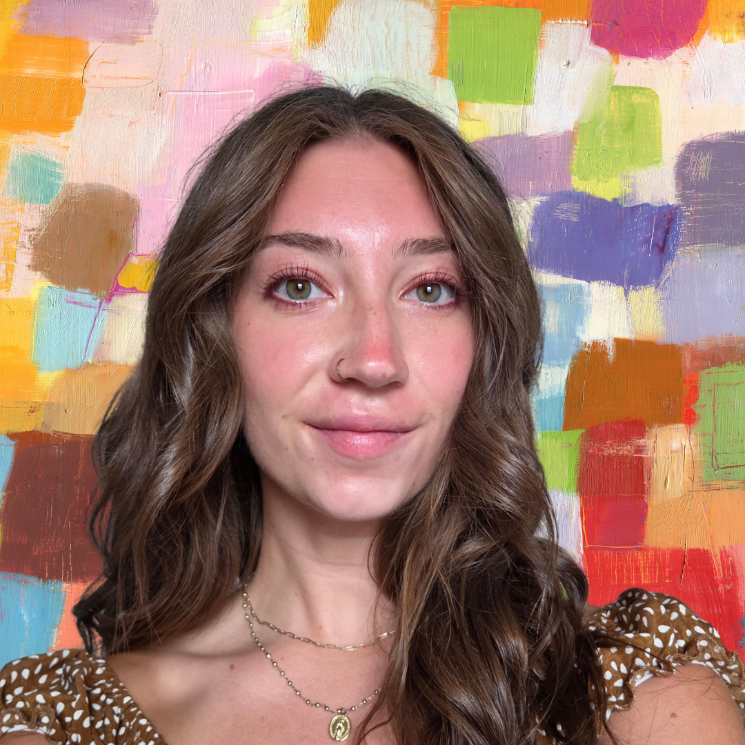 A young woman with wavy brown hair, hazel eyes, and light skin taking a selfie in front of a colorful abstract painting. She is wearing a brown polka dot top, gold necklaces, and has a small nose ring.