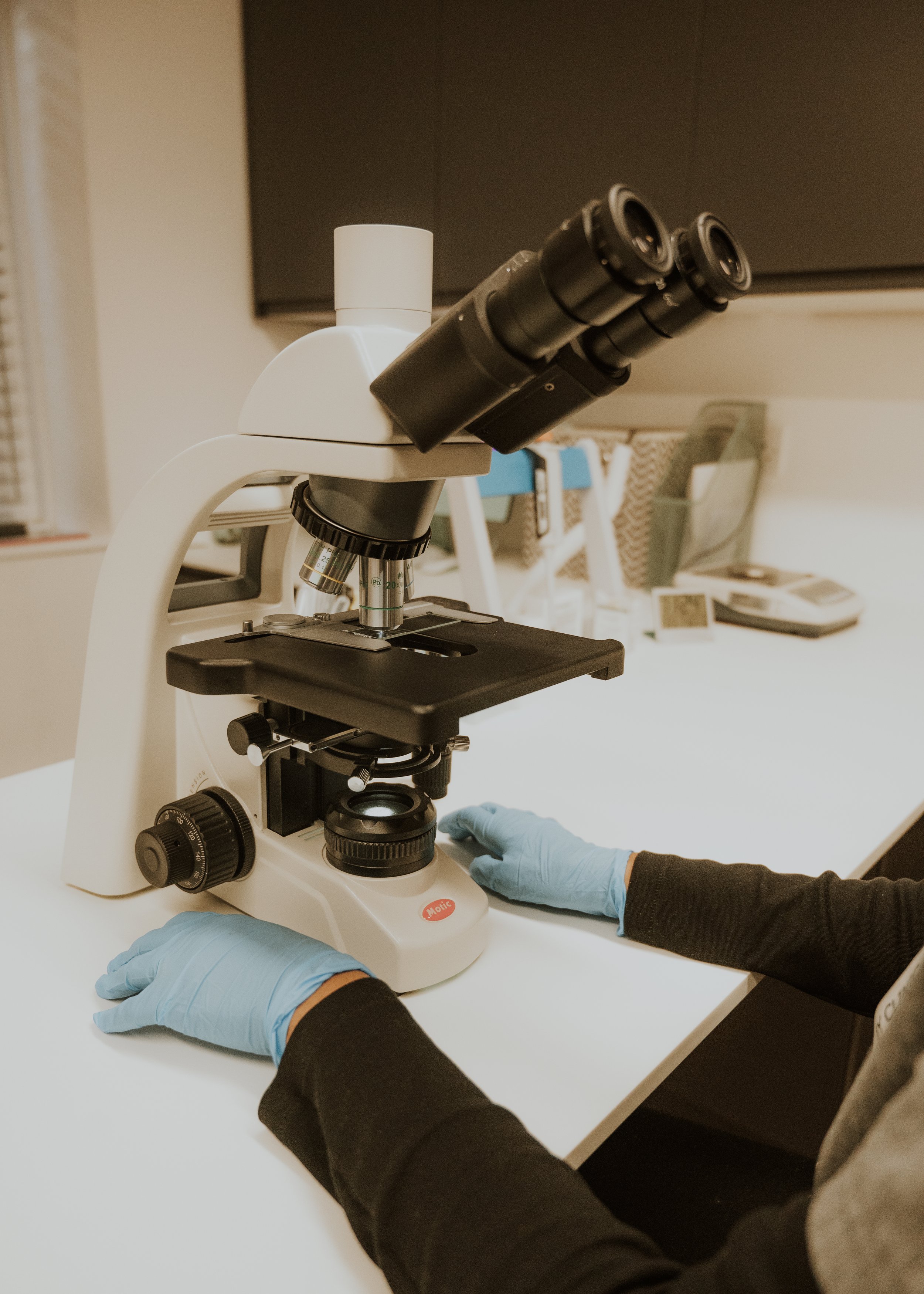 Close-up of a person wearing blue gloves using a compound microscope on a white laboratory table.