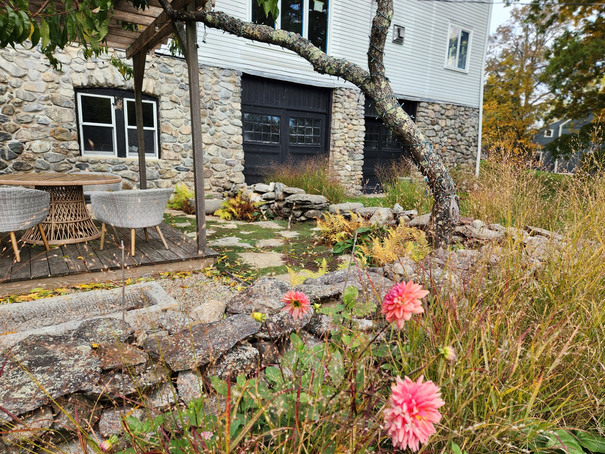 A backyard garden features a stone house with black garage doors, a wooden deck with patio furniture, a small rock garden, and flowering plants including pink flowers in the foreground. A tree with a trunk wrapped in string lights leans over the yard