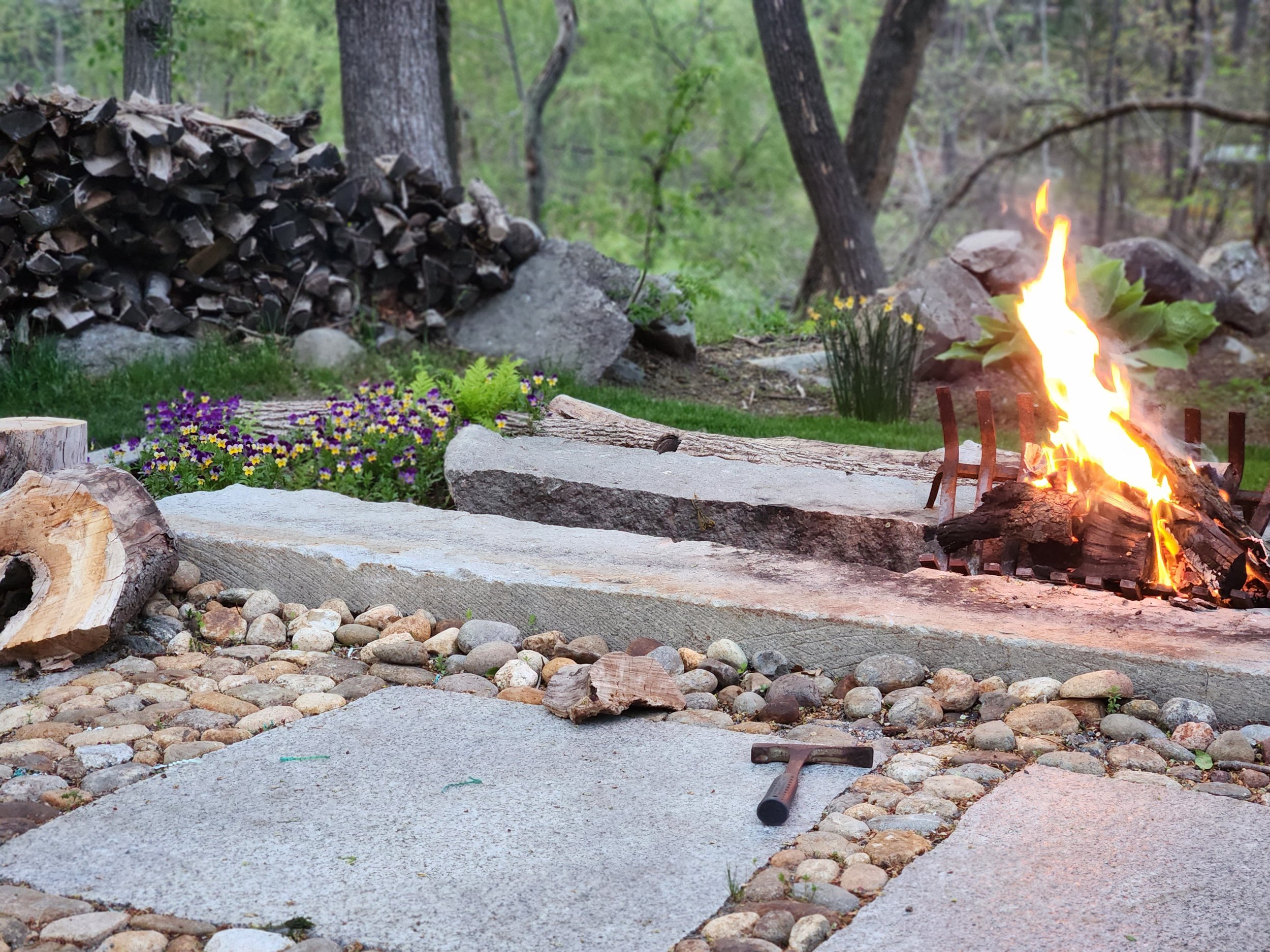 A backyard scene with a woodpile, fire pit with burning fire, rocks, and stone pathway surrounded by trees and greenery.