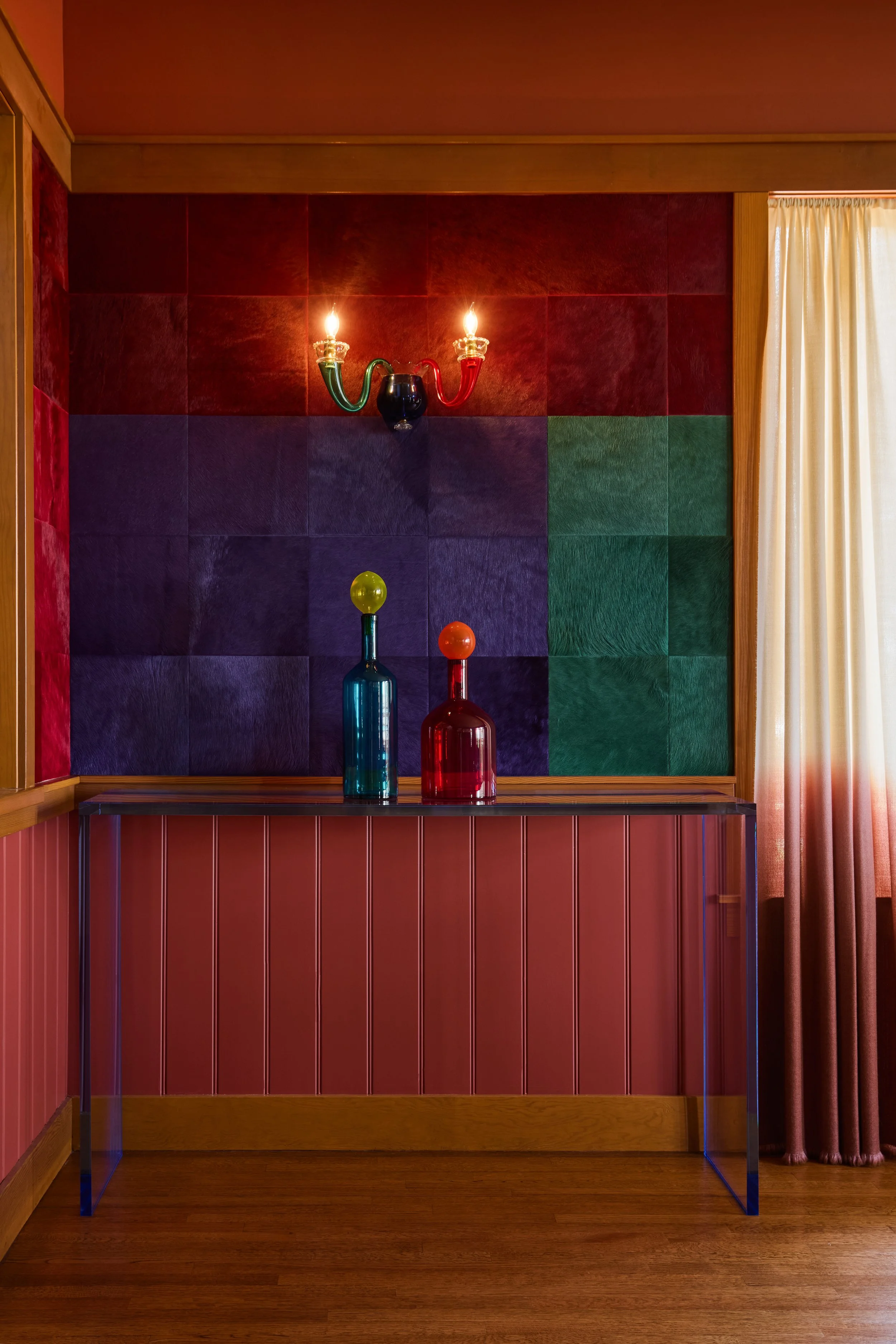 Eclectic interior design featuring a bold color-blocked wall in deep red, purple, and green tones. A sleek acrylic console table holds two sculptural glass vases in vibrant blue and red with playful spherical tops. A whimsical two-arm wall sconce add