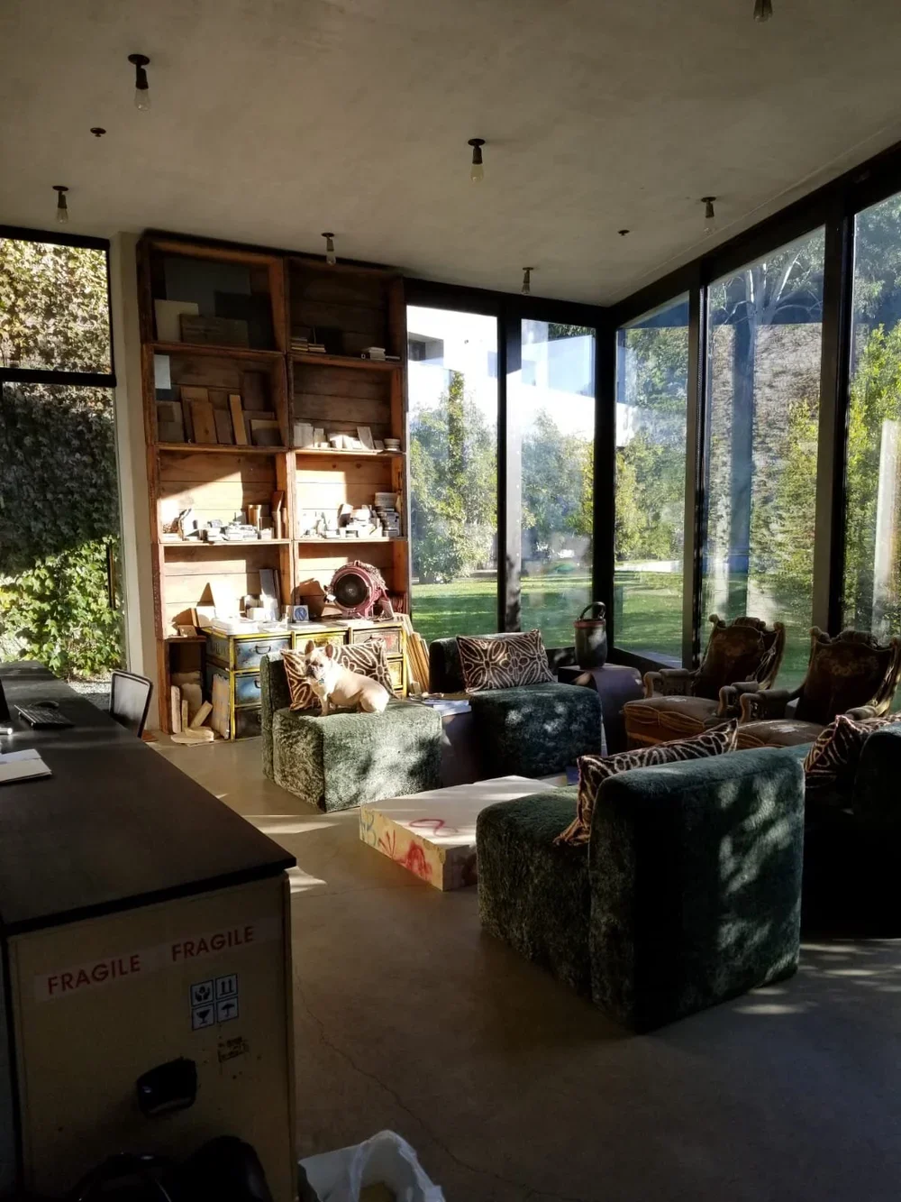 Sunlit creative studio interior featuring floor-to-ceiling glass walls, lush greenery views, vintage armchairs, velvet textured seating, and a rustic open shelving unit filled with art supplies and materials. This eclectic lounge space combines bohem