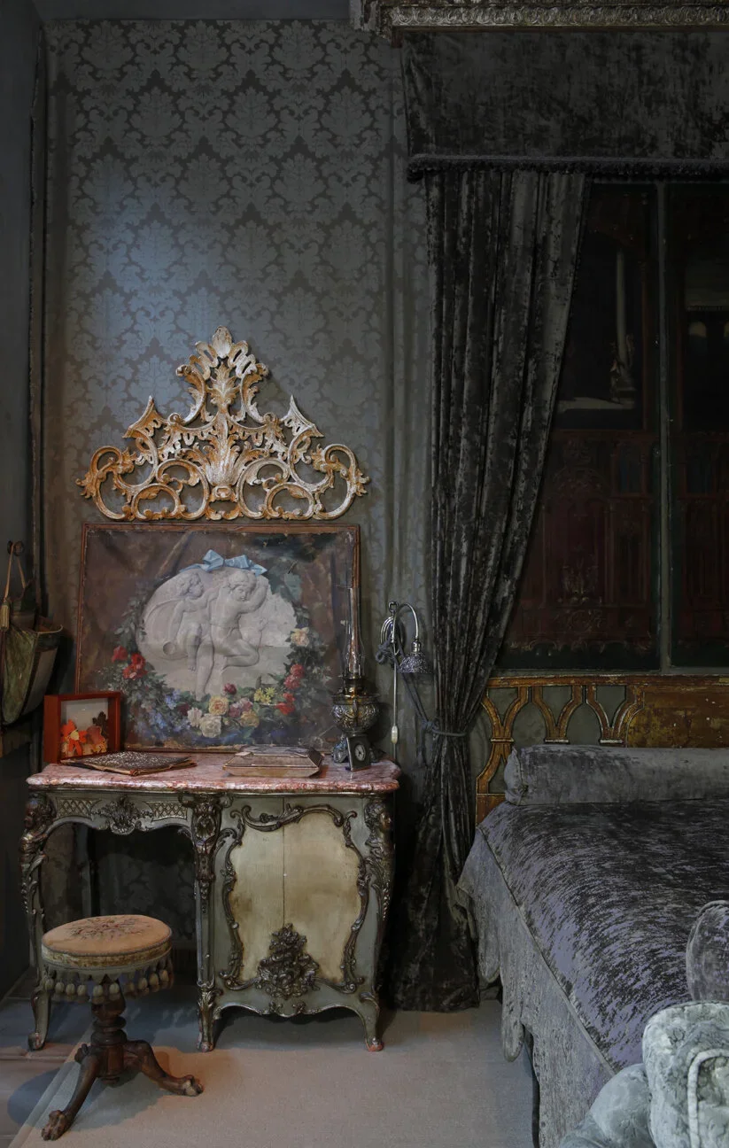 Luxurious vintage bedroom interior with ornate antique furniture, velvet drapes, and a classic carved dressing table topped with artwork and a decorative lamp. The rich textures and intricate details create a romantic, old-world ambiance that exudes